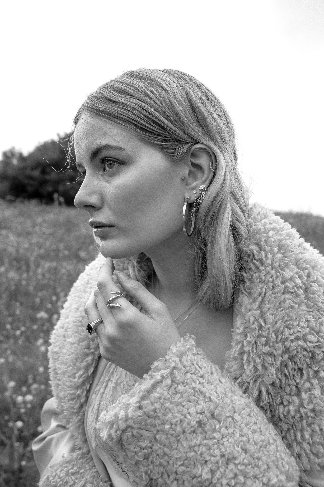 Black and white portrait of a woman with blond hair wearing hoop earrings and a fluffy coat, standing in a field.
