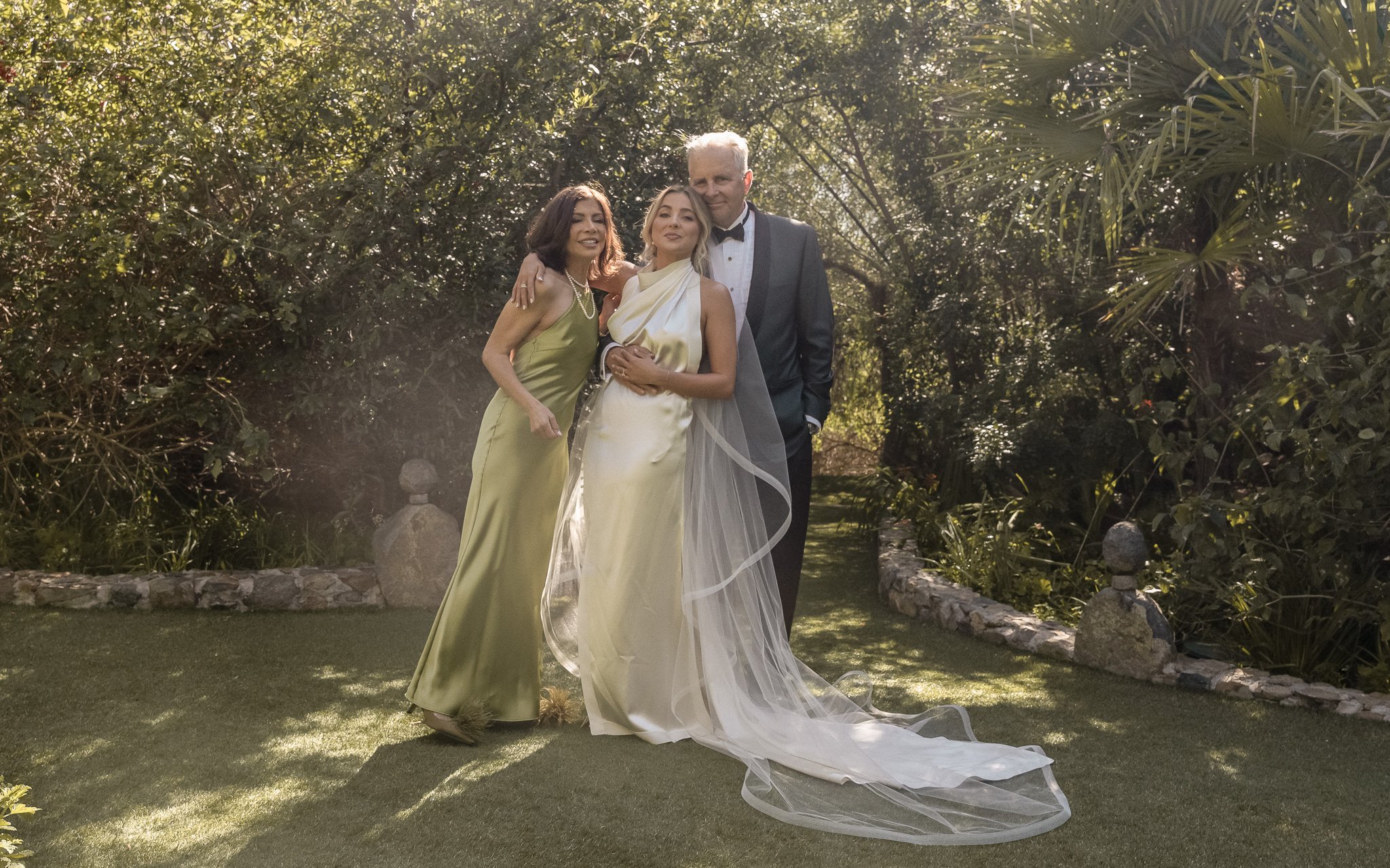 Three people pose outdoors in formal attire; one in a wedding dress, flanked by two individuals in formal wear, in a lush garden setting.