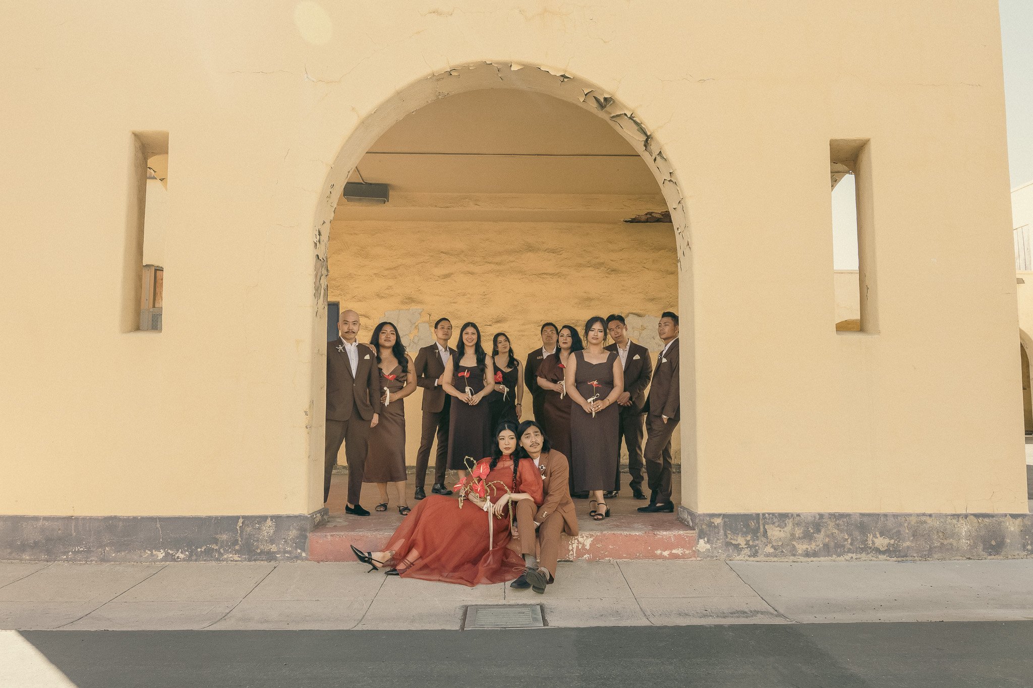 A group of people in formal attire posing under an archway. Two individuals are sitting on the ground in front, while the others stand behind them, arranged in a semi-circle under the arch. The background features a textured beige wall.