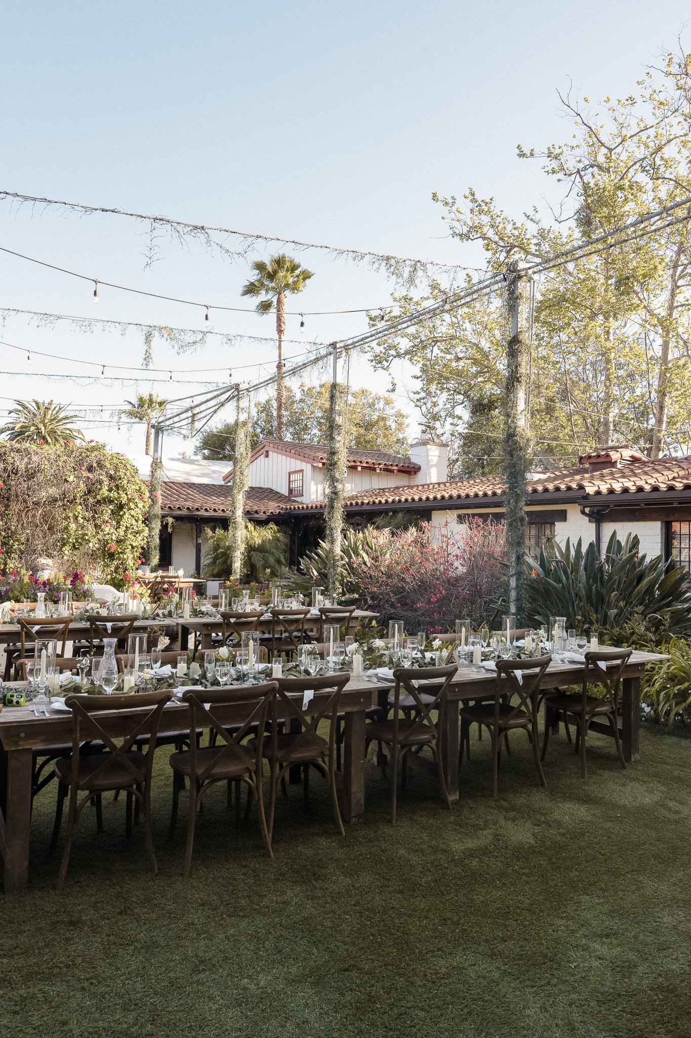 Outdoor garden dining setup with wooden tables and chairs, decorated with candles and greenery, surrounded by plants and trees, under string lights.
