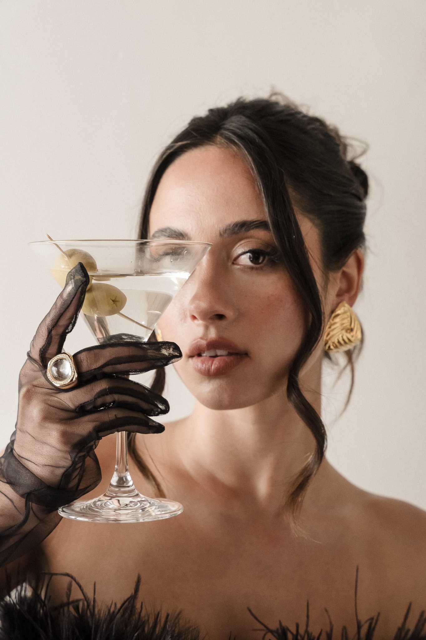Person with elegant hairstyle holding a martini glass with olives, wearing a black lace glove and large gold earring, with neutral background.