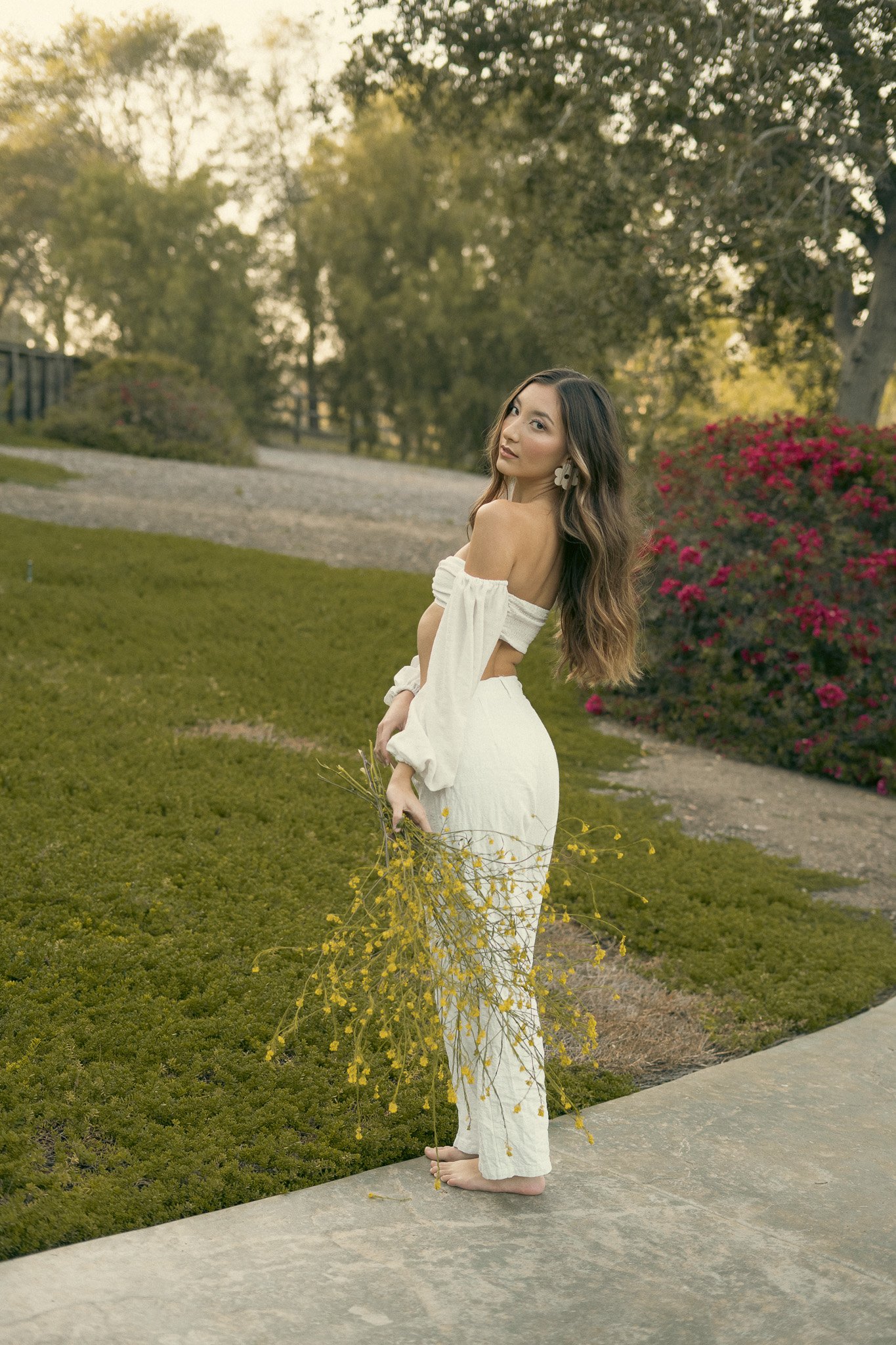 A woman in a white off-shoulder dress holding yellow flowers, standing barefoot on a garden path with greenery and blooming bushes in the background.