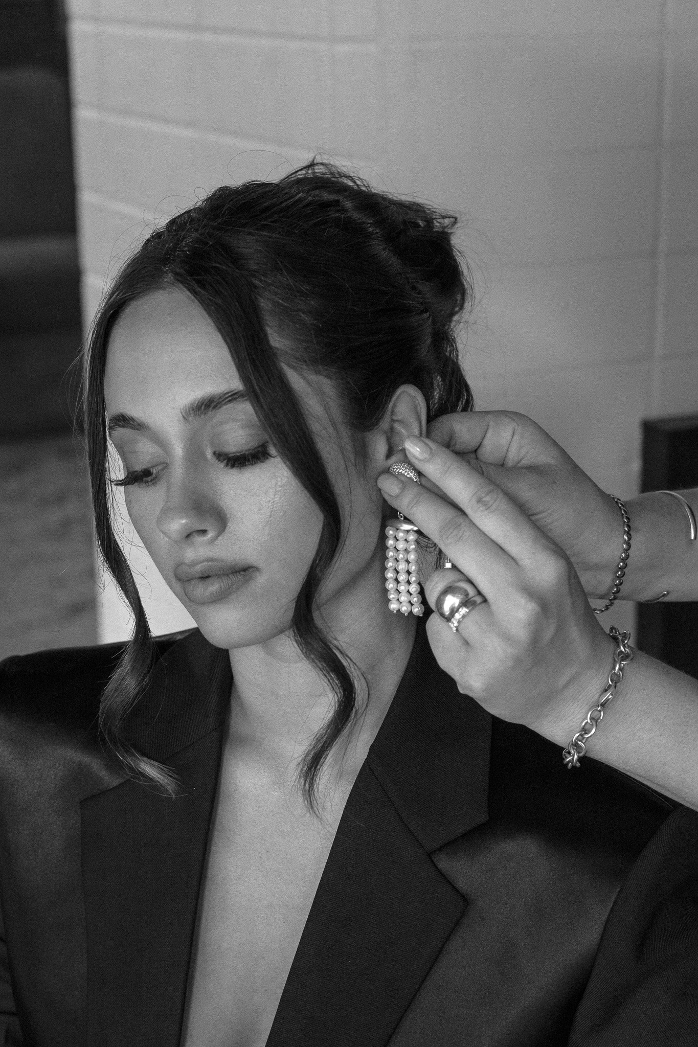 Woman wearing a black blazer having an earring with dangling pearls put on her by a hand.