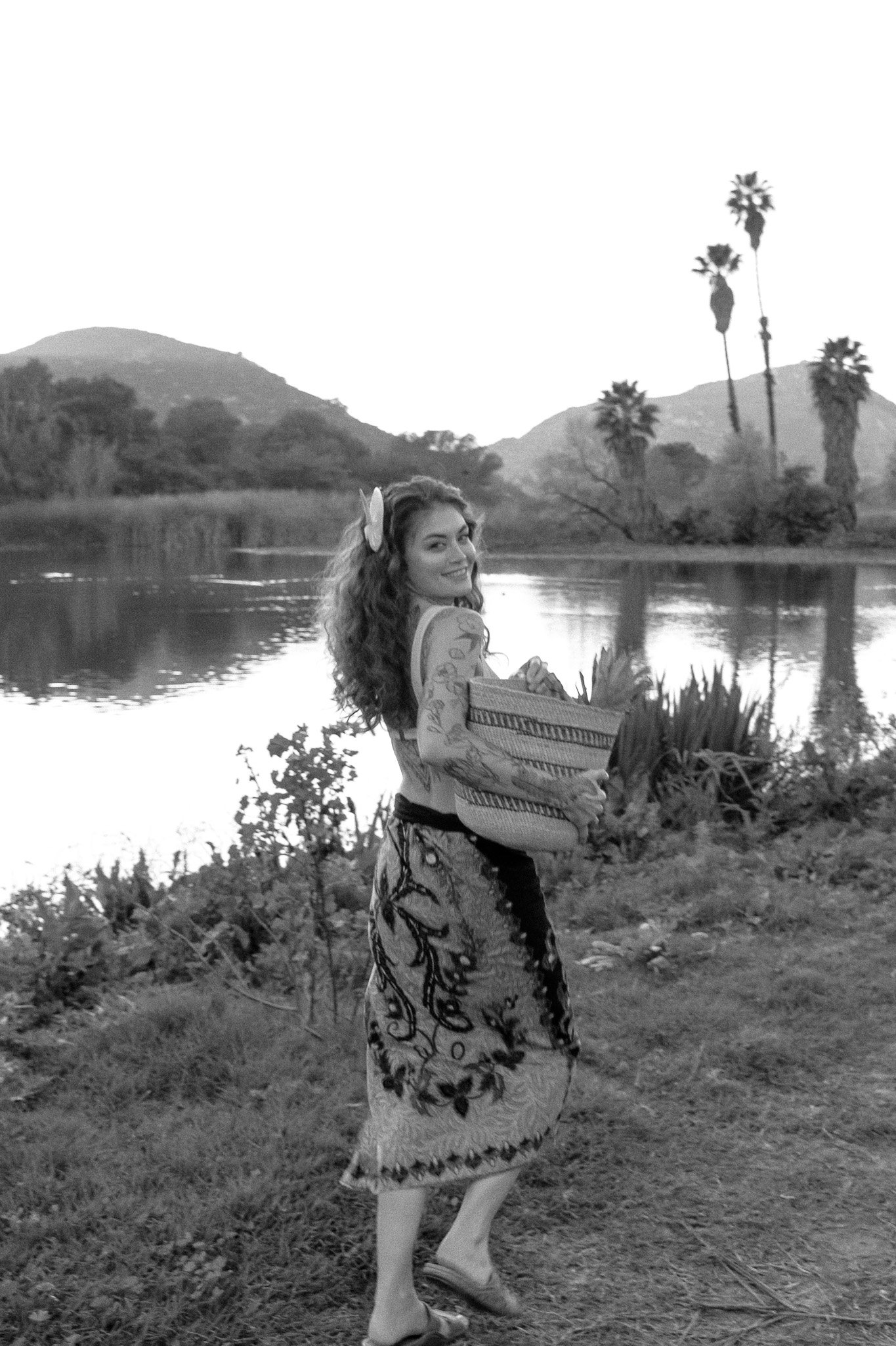 Woman with tattoos holding a woven basket near a lake, palm trees and mountains in the background, black and white.