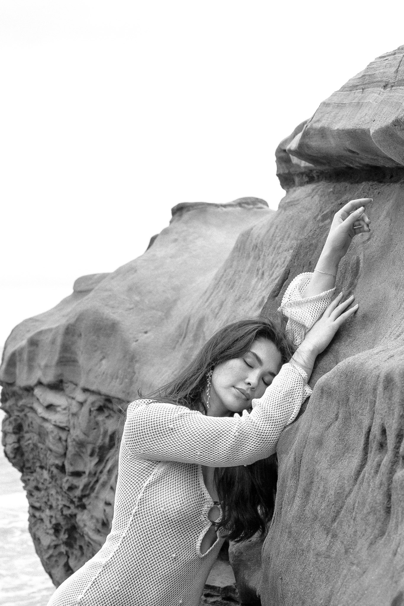 Black and white photo of a woman with long hair leaning against large rock formations, eyes closed, wearing a netted outfit and earrings.