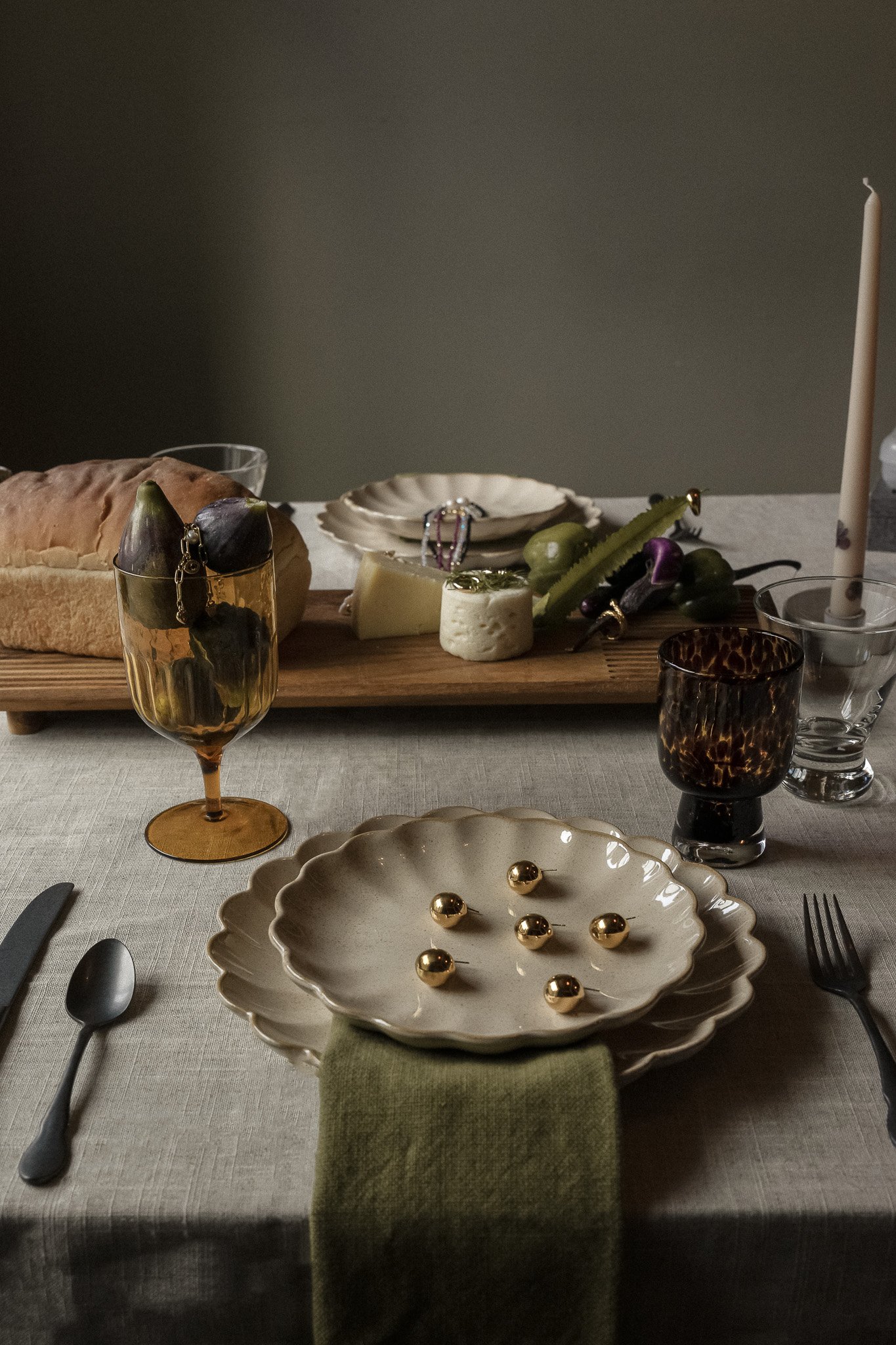 Elegant table setting with ceramic plates, cutlery, amber glass with figs, bread loaf, cheese, assorted vegetables on a wooden board, decorative golden spheres on plates, and a tall candle.