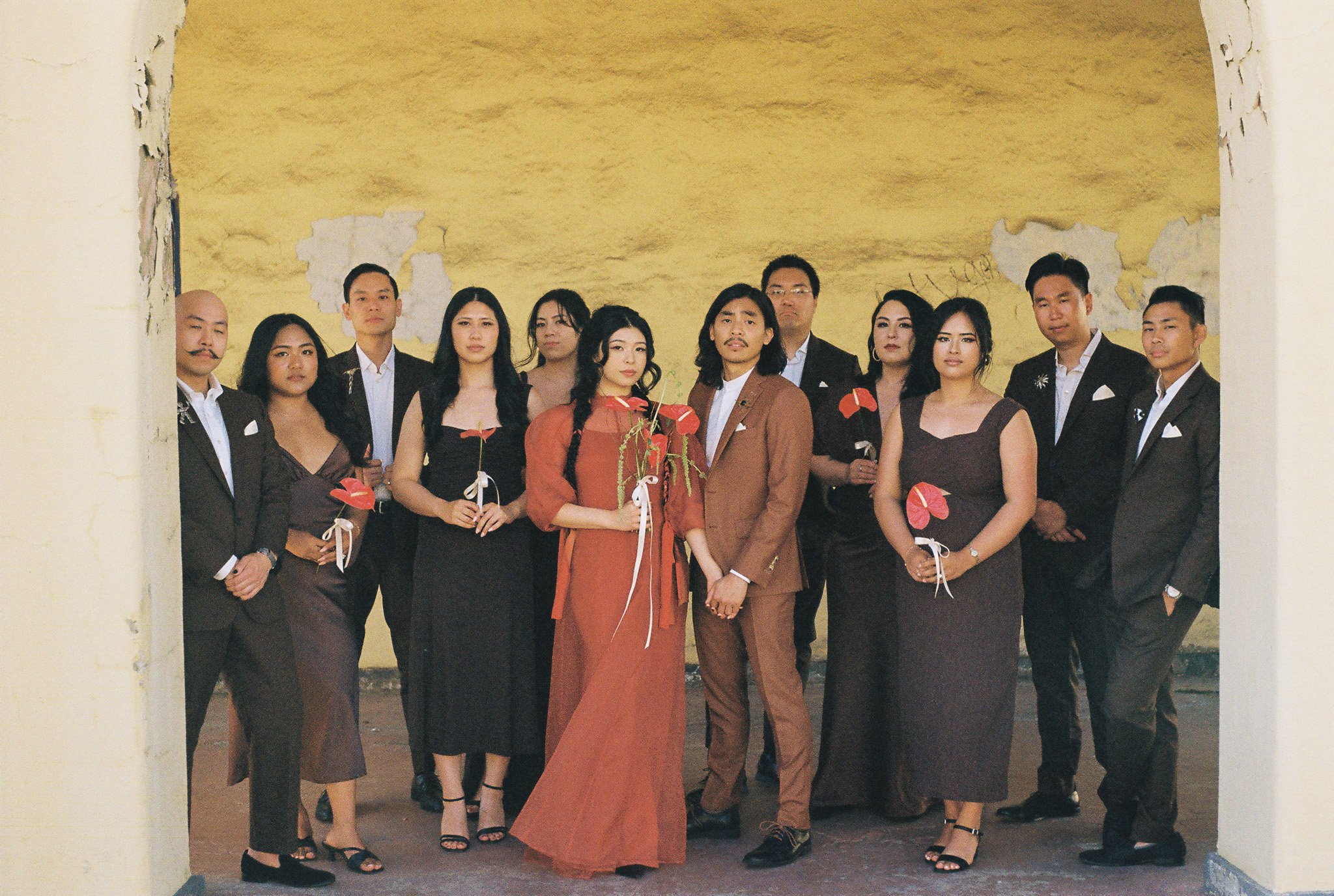A group of formally dressed people standing together, some holding red flowers, in front of a textured yellow wall.