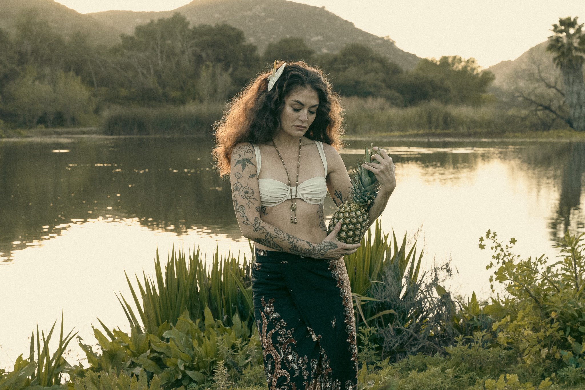 Woman with tattoos holding a pineapple in a natural outdoor setting.