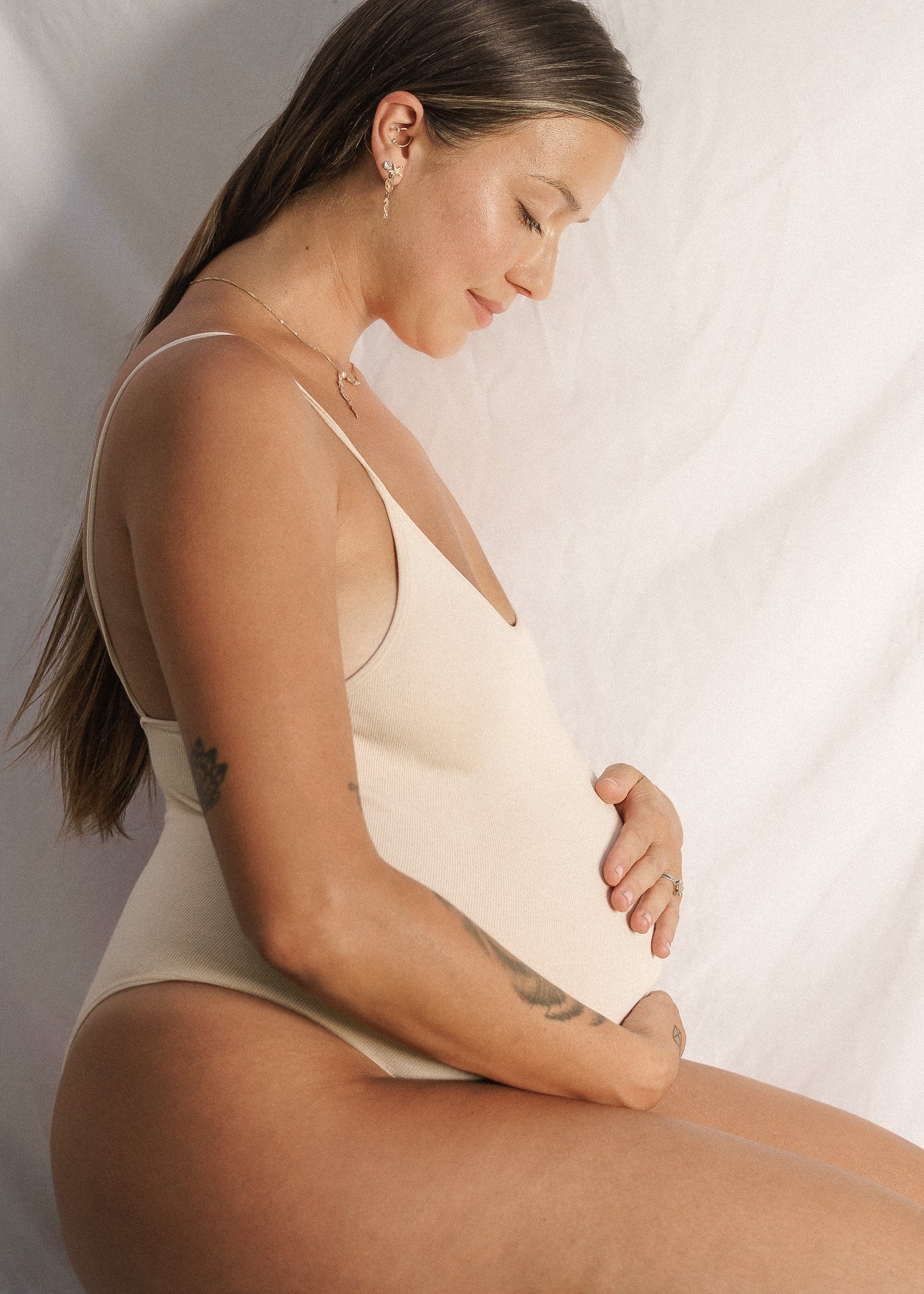 Pregnant woman in beige clothing holding her belly, sitting against a light background.