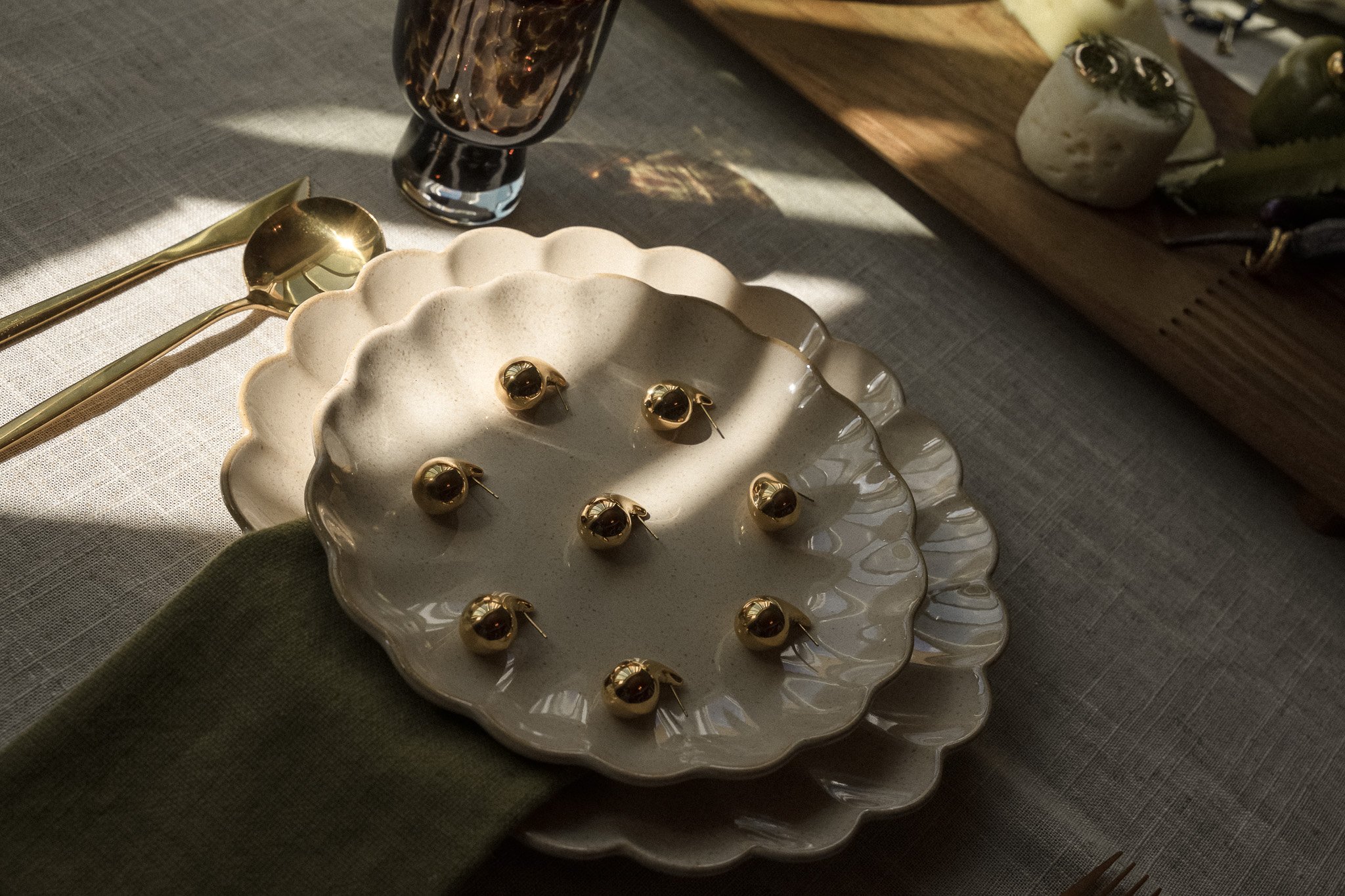 Elegant table setting with stacked scalloped plates topped with gold earrings, gold cutlery, a dark glass cup, and a wooden board on a beige tablecloth.