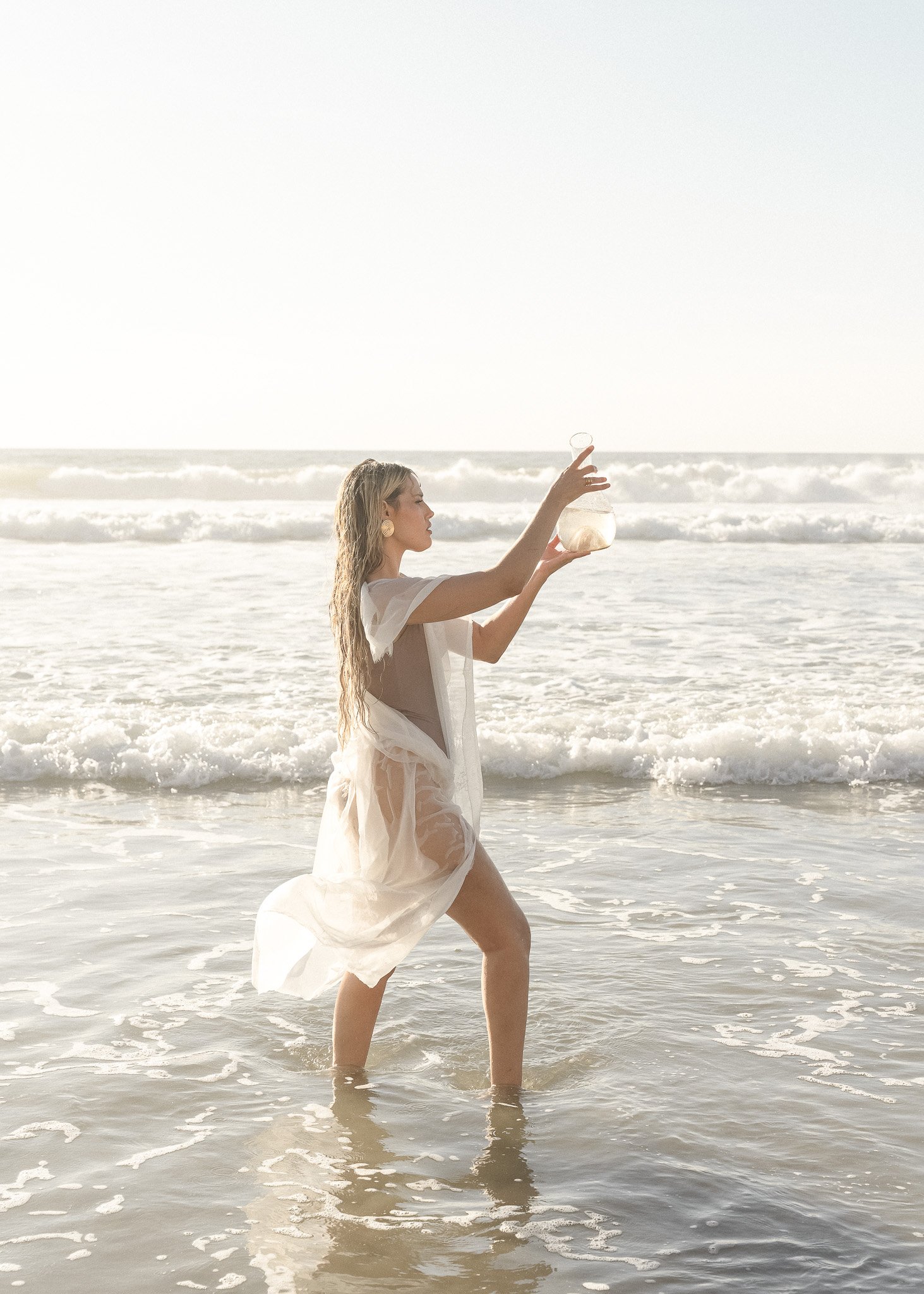 Woman in a sheer white dress holding a transparent orb, standing in shallow ocean water with waves and a clear sky in the background.