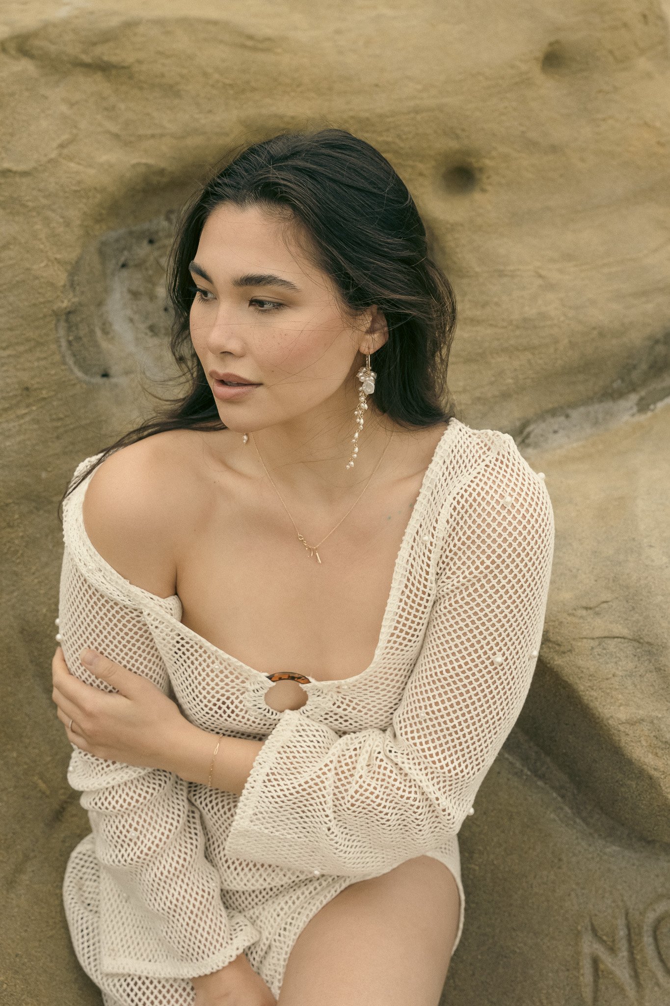 Woman in white mesh outfit sitting on a rock, looking sideways, with long earrings and natural background.