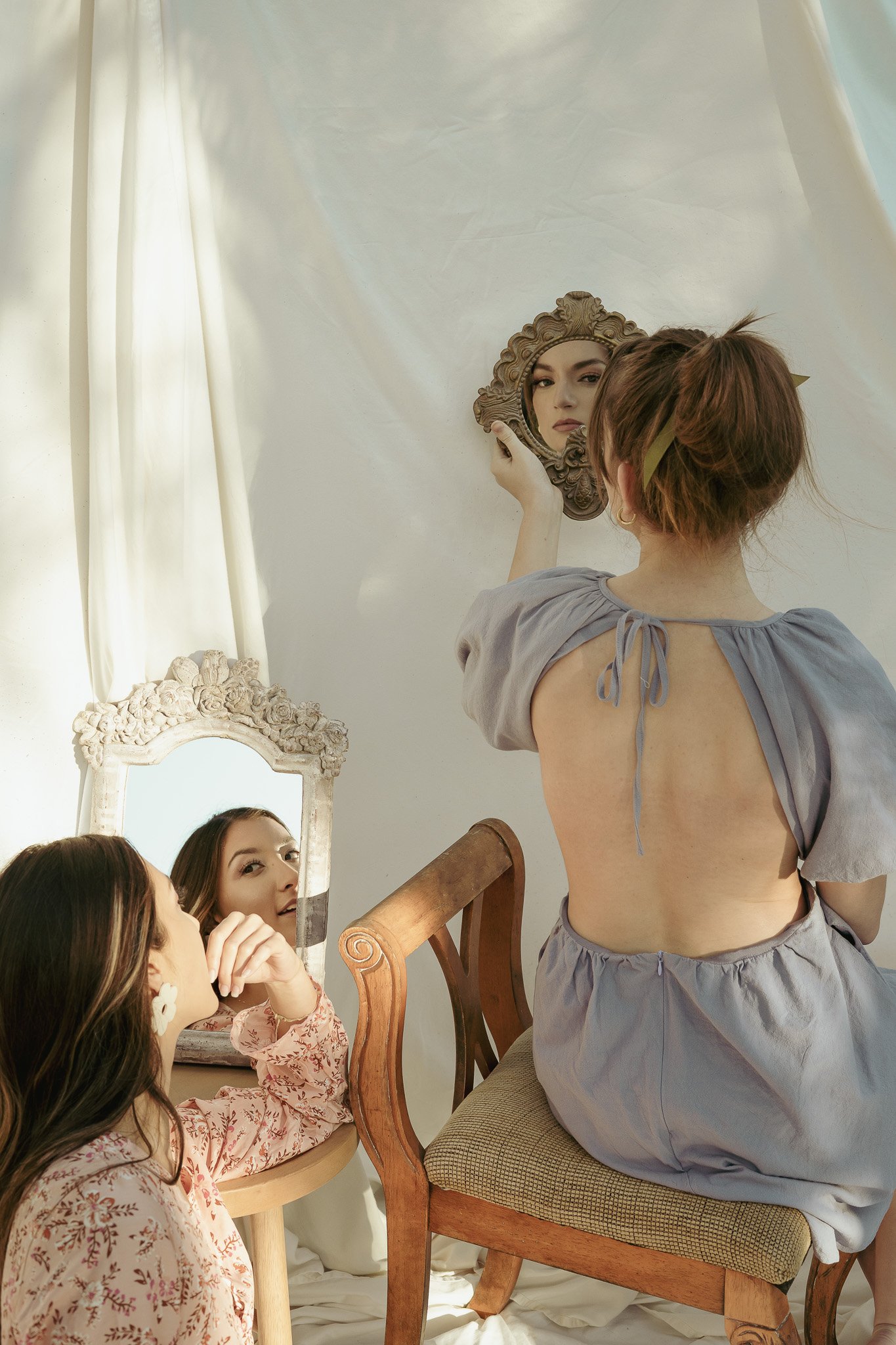 Two women sit indoors, one with a backless dress looking into an ornate hand-held mirror, the other leaning against a wooden chair gazing into a tabletop mirror. Soft light filters through curtains.