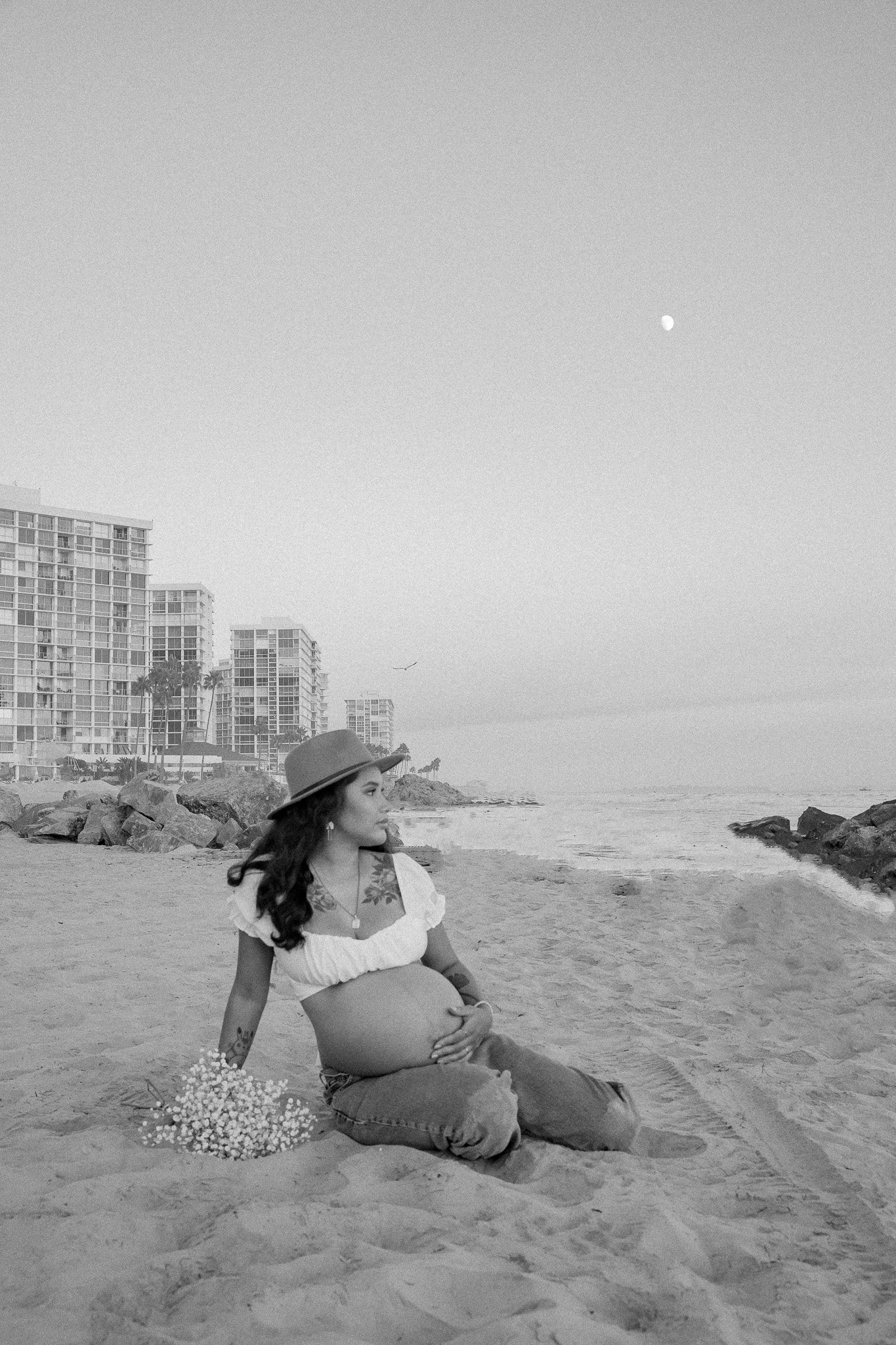 Pregnant woman sitting on a beach, wearing a hat and white blouse, holding her belly, with buildings and the ocean in the background.