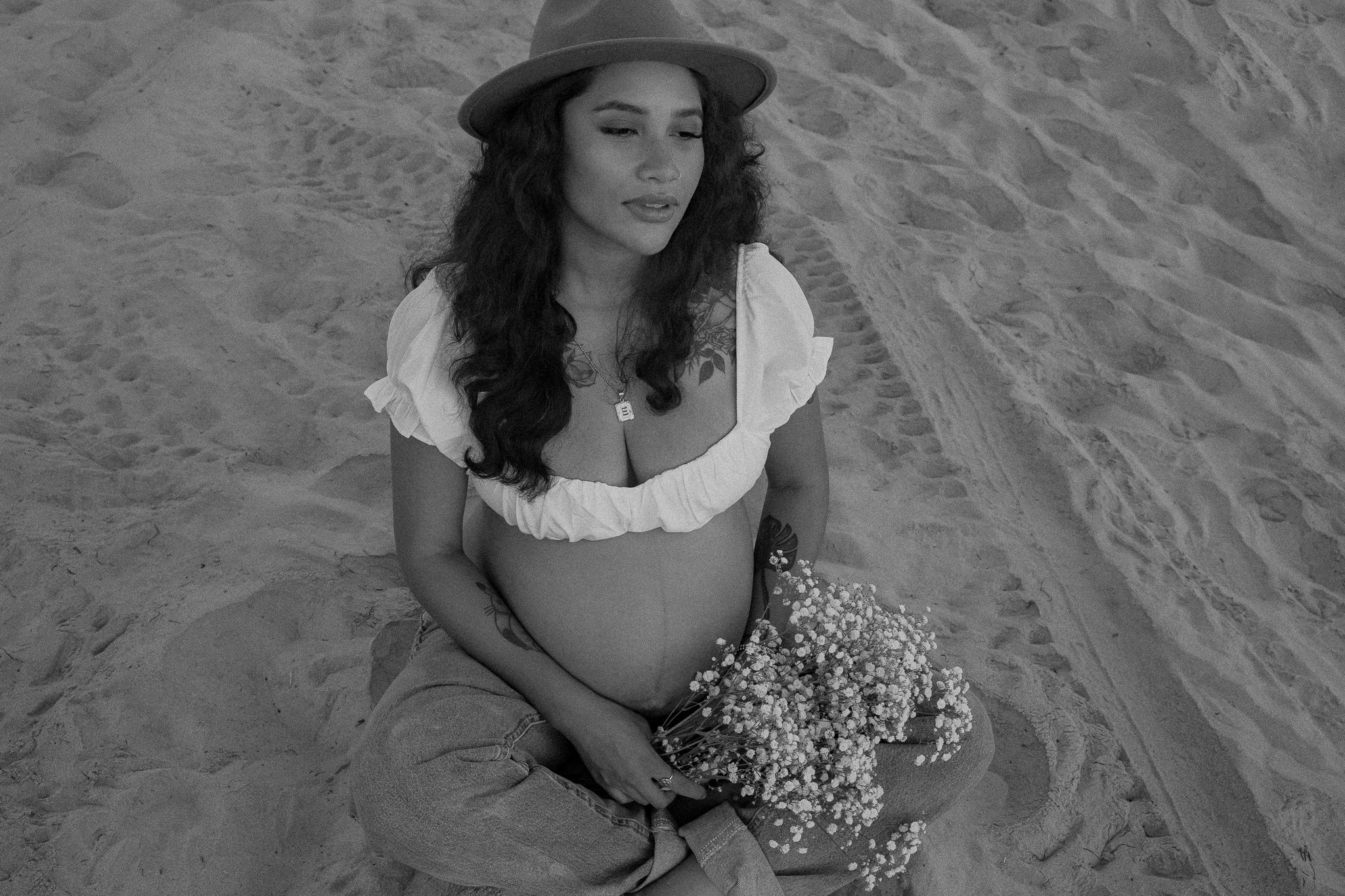 Pregnant woman sitting on sand holding white flowers, wearing a hat and a white top, black and white photograph.