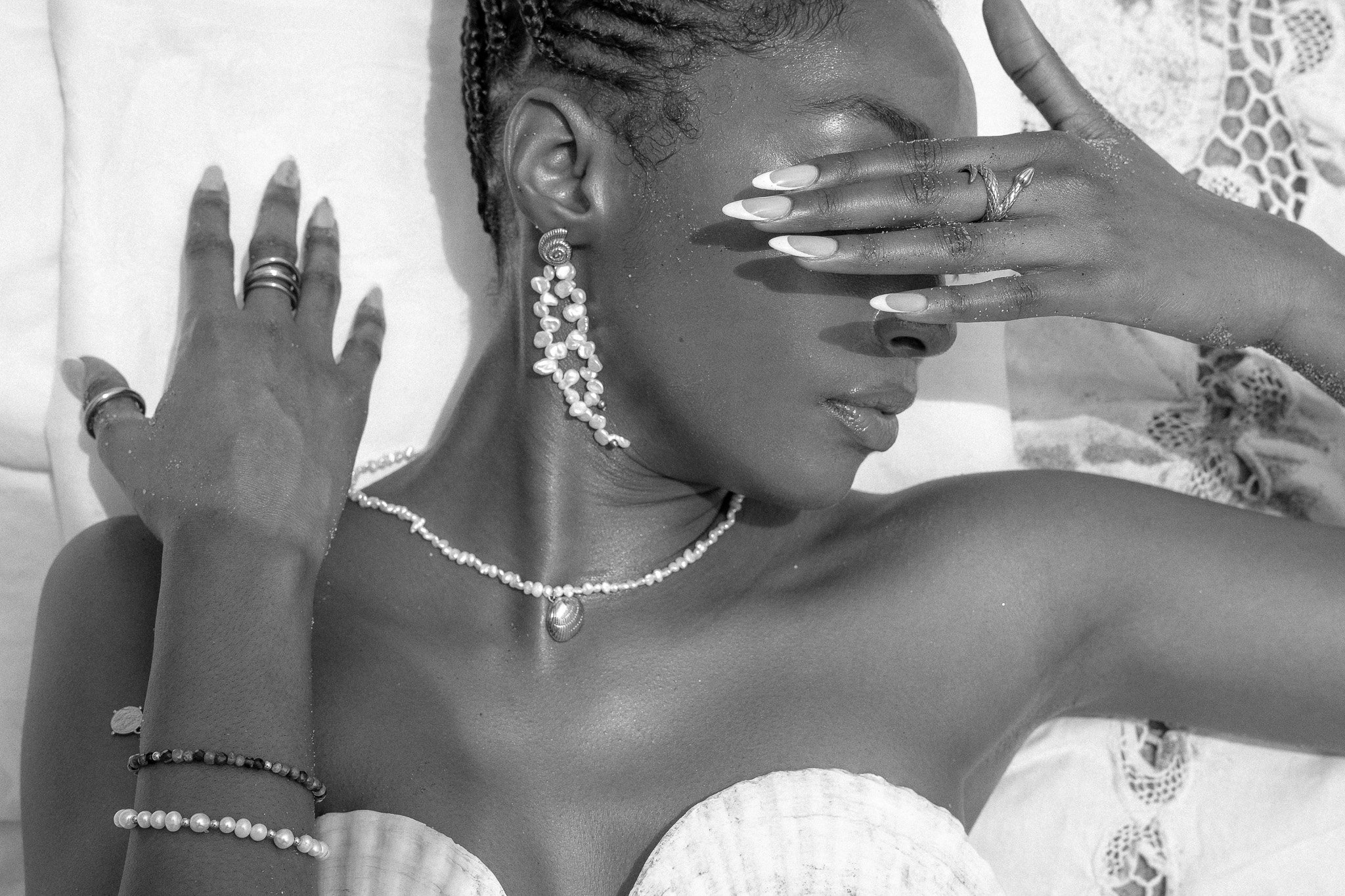 Black and white image of a woman with braided hair wearing pearl earrings and necklace, raising her hand to cover her eyes, with rings and bracelets on her fingers.
