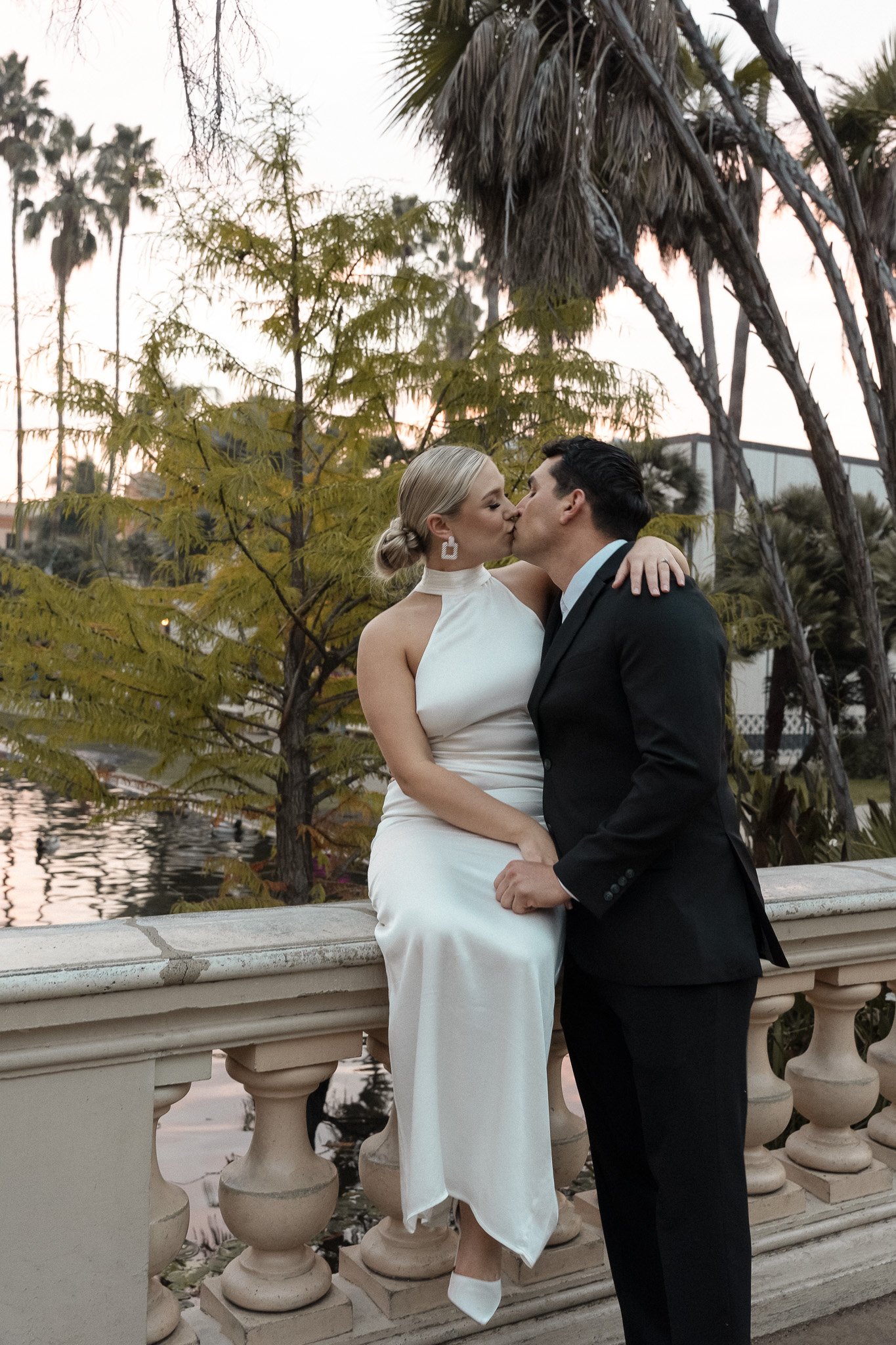 Couple in formal attire kissing on a stone railing with palm trees and a pond in the background.
