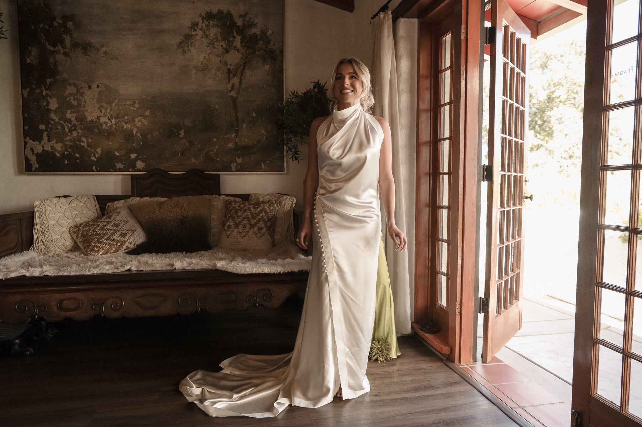 Woman in a white satin dress standing indoors near a wooden door, next to a couch with decorative pillows and a nature painting on the wall.