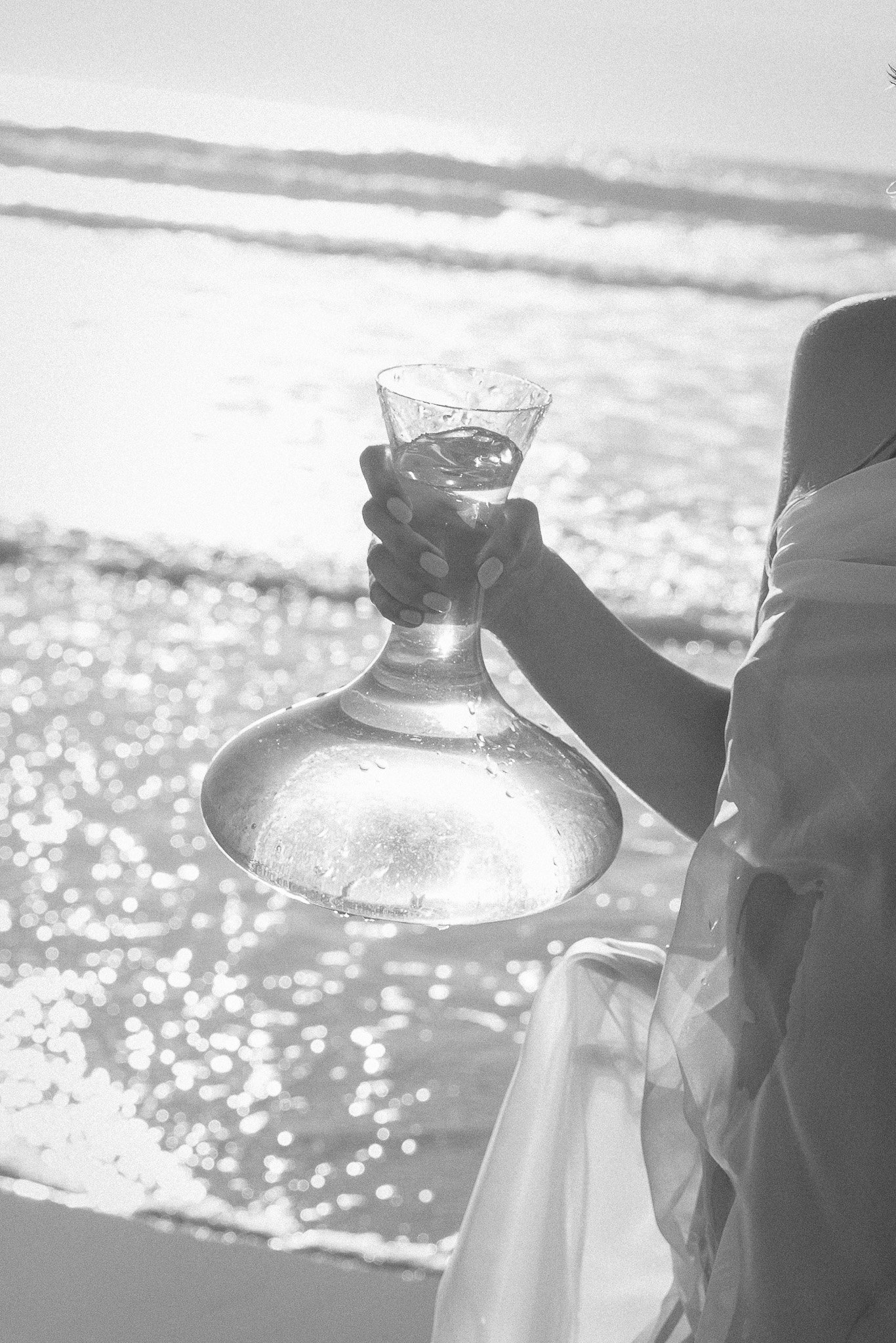 Black and white image of a person holding a glass decanter with water by the ocean