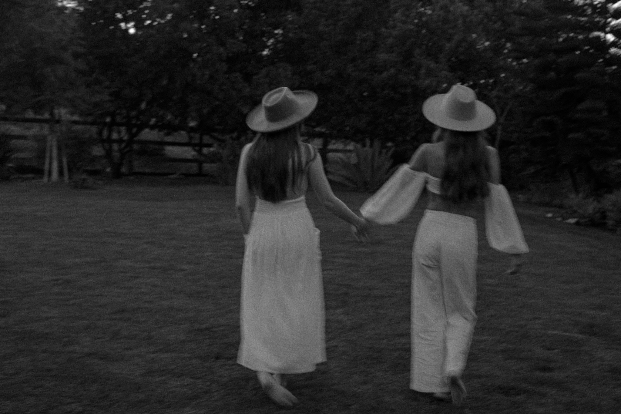 Two women in hats and long dresses walking hand in hand on grass, outdoors in a natural setting.