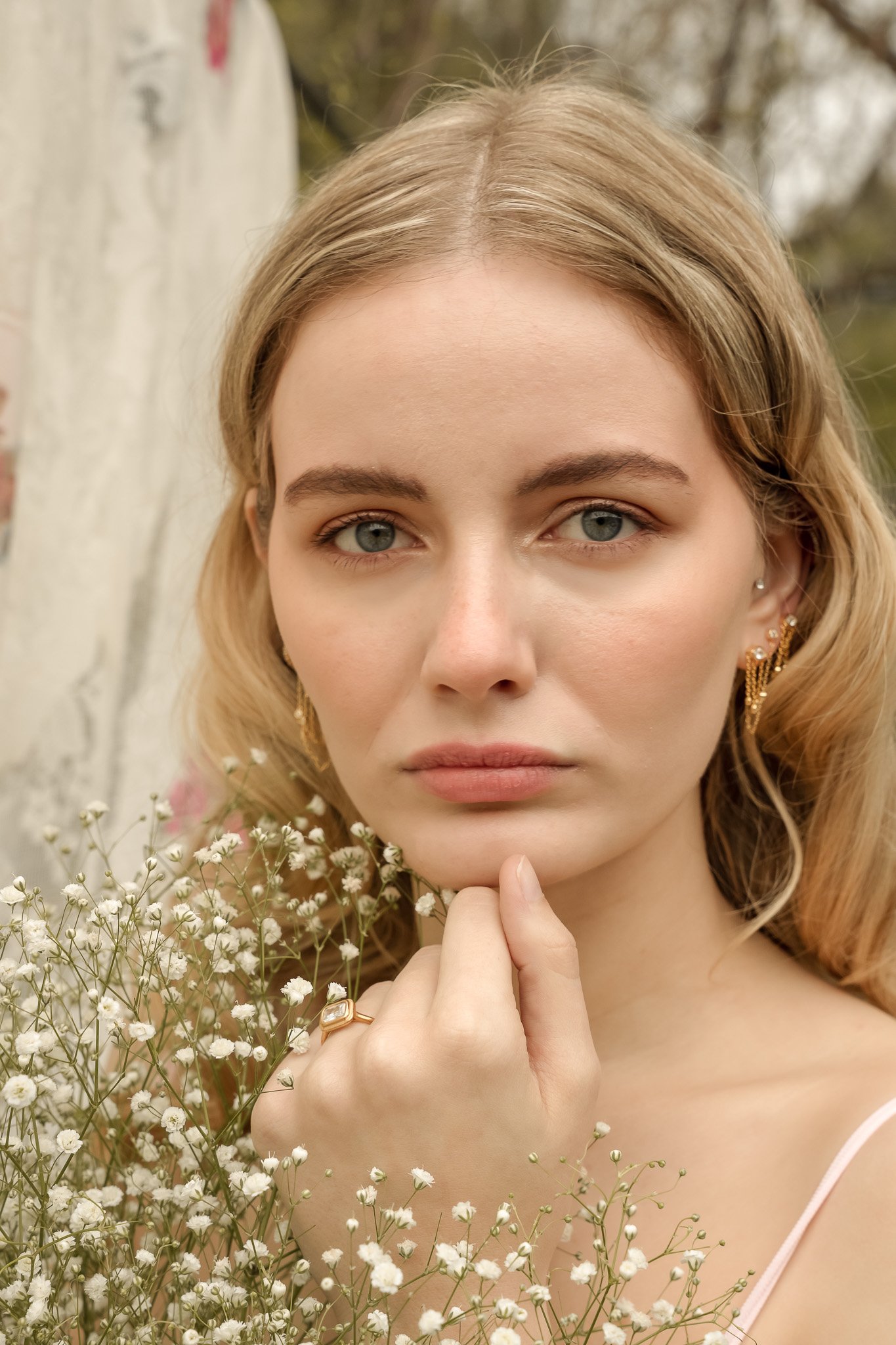Young woman with blond hair and gold earrings, surrounded by white flowers, looking at the camera with a soft expression.