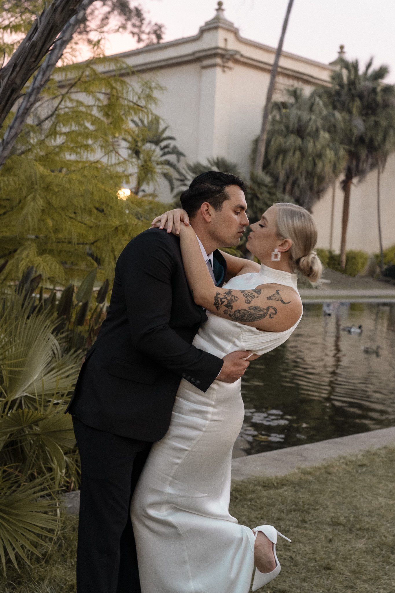 Bride and groom sharing a kiss outdoors by a pond, with a building and palm trees in the background.