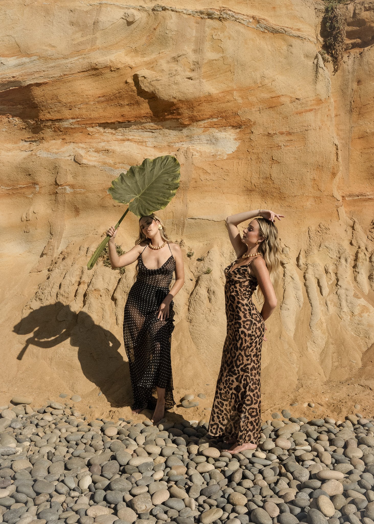 Two women standing against a rocky sand-colored wall, one holding a large leaf, wearing fashionable dresses, on a rocky beach.