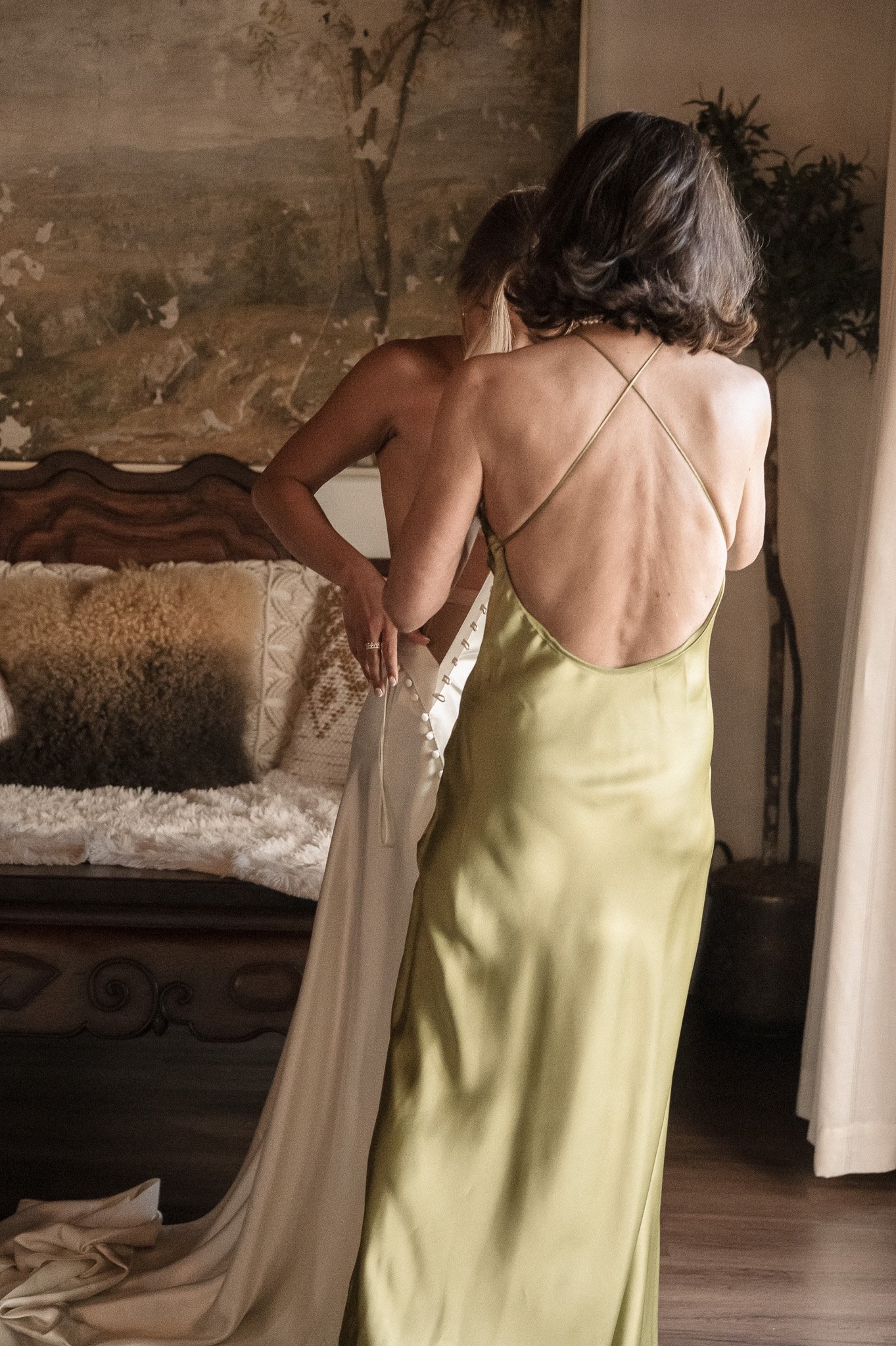 A woman in a green dress adjusts another woman's white dress in a cozy, elegantly decorated room.