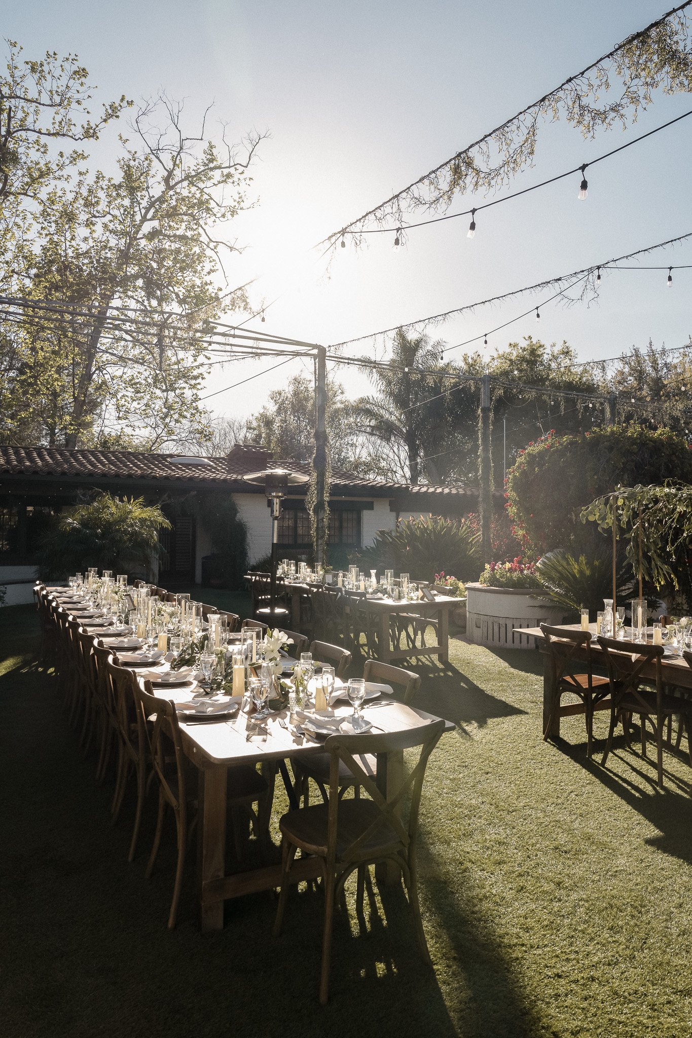 Outdoor wedding reception setup with long tables, chairs, place settings, candles, hanging string lights, and greenery, in a garden setting.