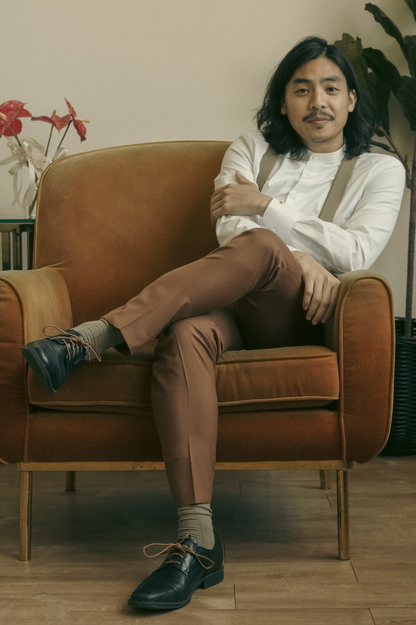 A man with long hair and a mustache sitting in a brown armchair, wearing a white shirt, suspenders, brown pants, and black shoes, with legs crossed. Indoor plant and red flowers in the background.