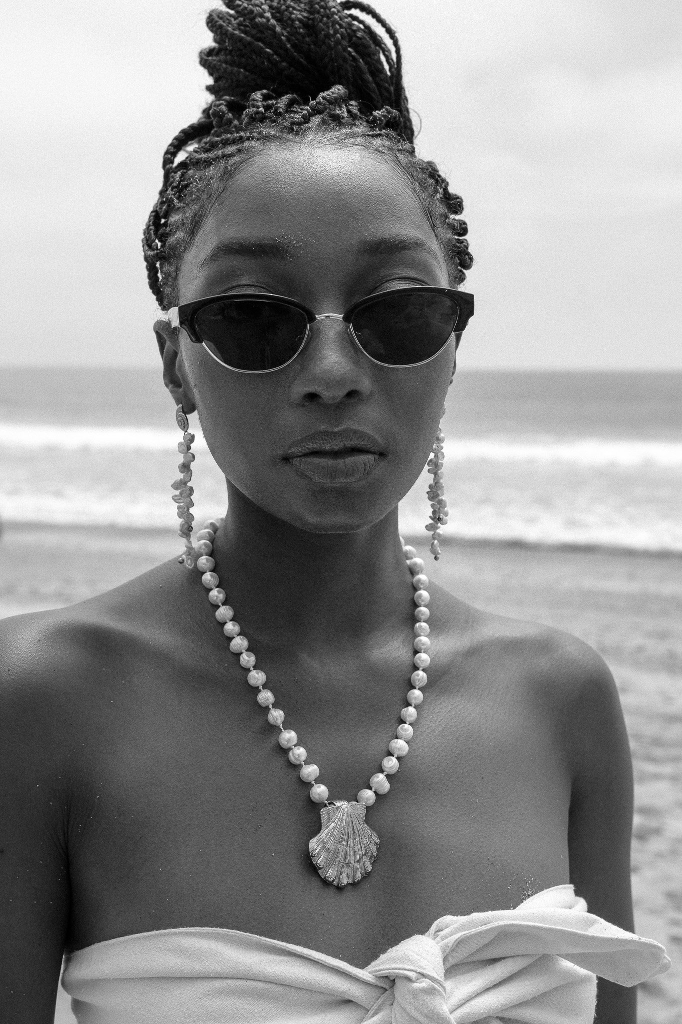 Black and white photo of a person wearing sunglasses and braided hair, with pearl jewelry, on a beach.
