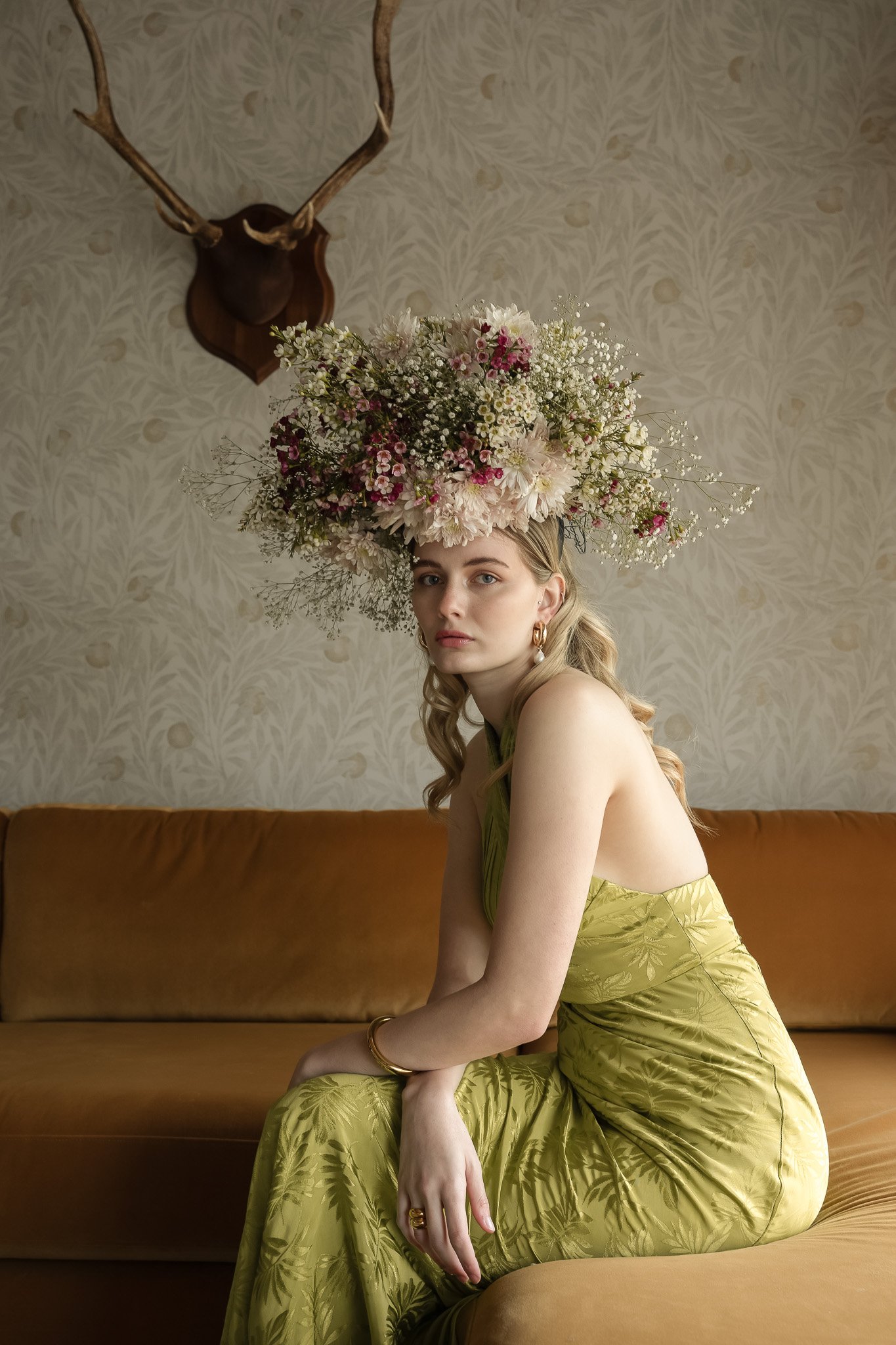 Woman in a green dress with a large floral headpiece sitting on a mustard sofa, antler decor on the wall.