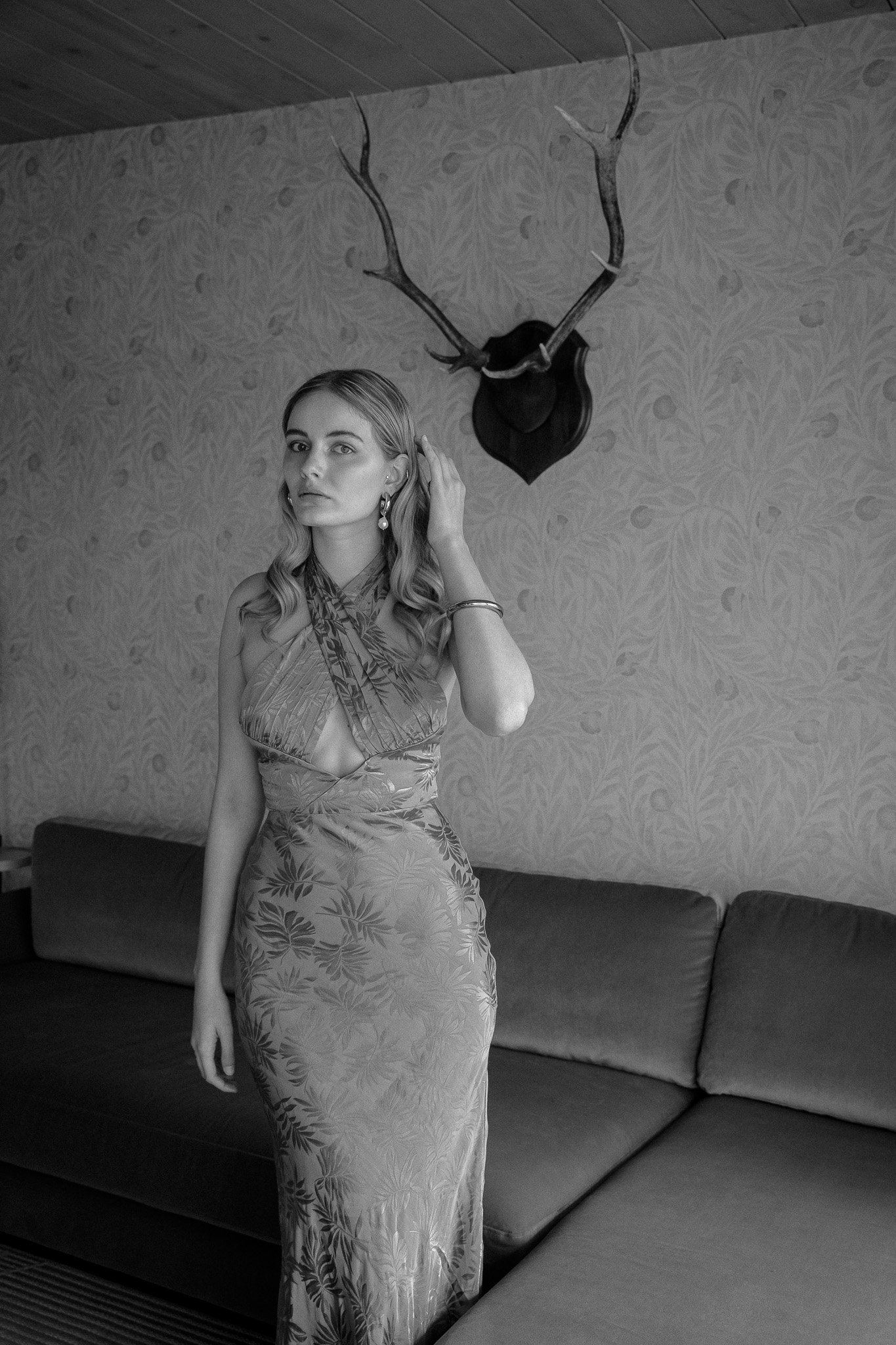 Black and white photo of a woman in a patterned dress standing indoors, with antlers mounted on the wall behind her.