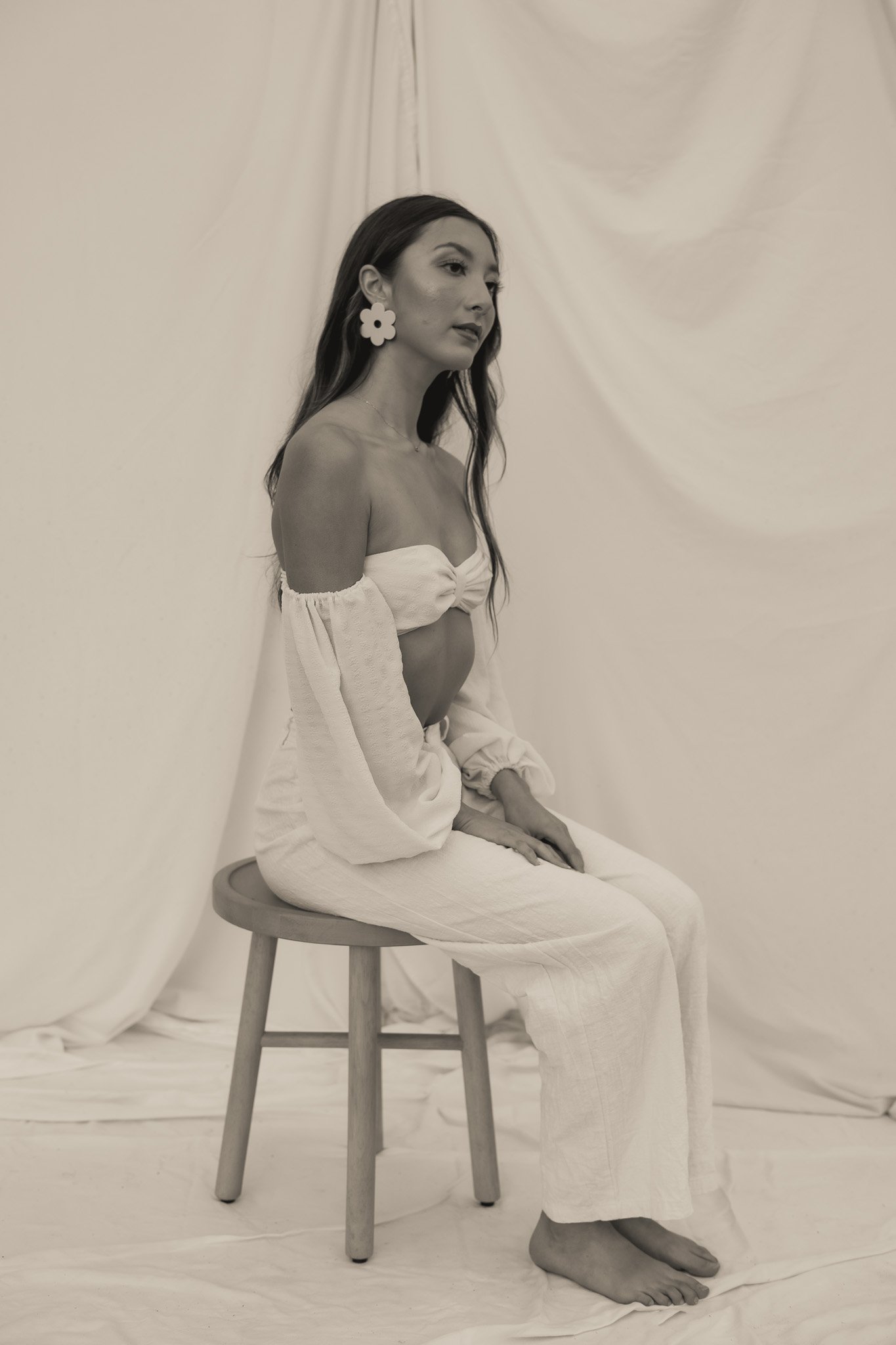 Woman in off-shoulder top and wide-leg pants sitting on a stool, wearing large floral earrings, against a draped fabric background.
