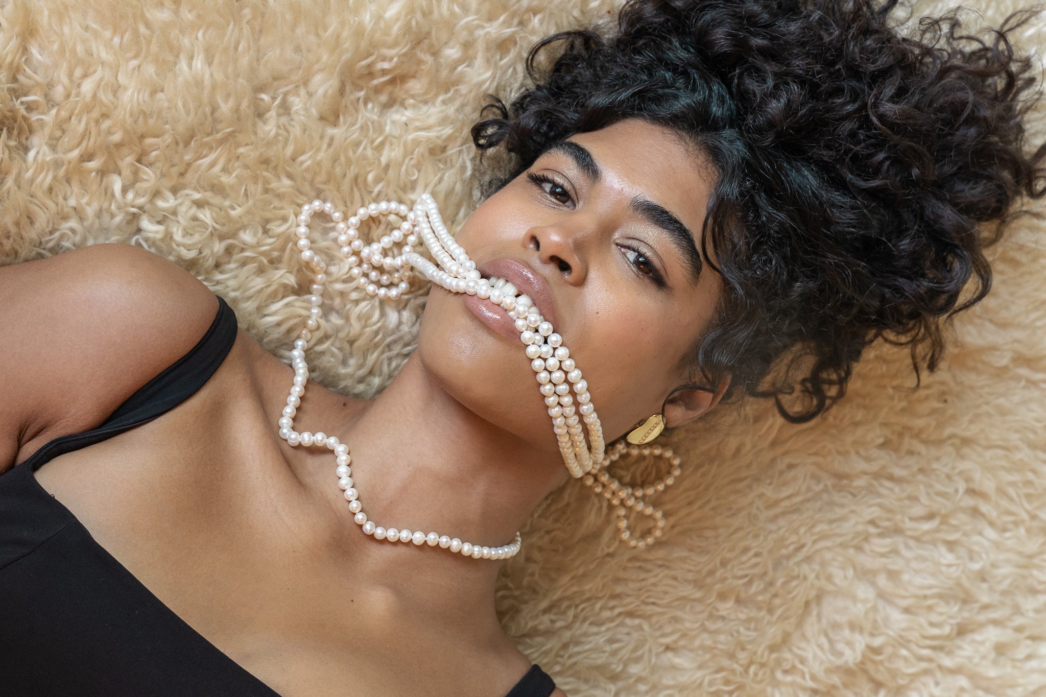 Woman with curly hair lying on a fluffy surface, draped with pearl necklaces, wearing a black top.