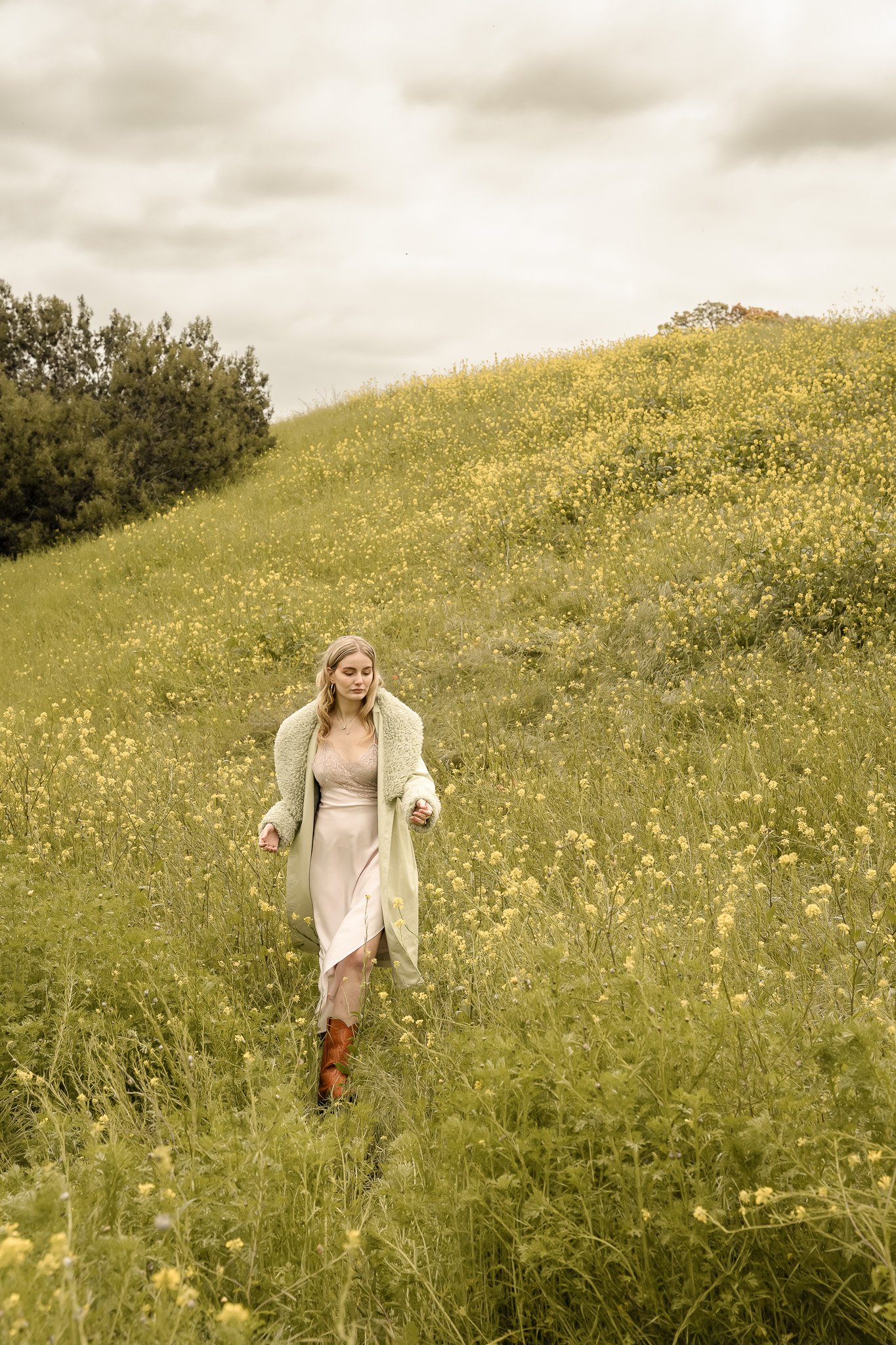 Woman in light green coat and white dress walking through a field of yellow wildflowers with overcast sky.