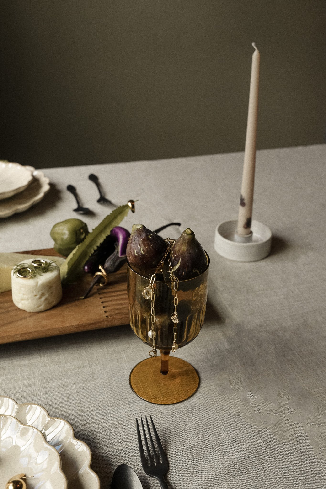 Elegant table setting with figs in a gold goblet, assorted cheese and vegetables on a wooden board, black cutlery, stacked white plates, and a tall candle on a gray tablecloth.