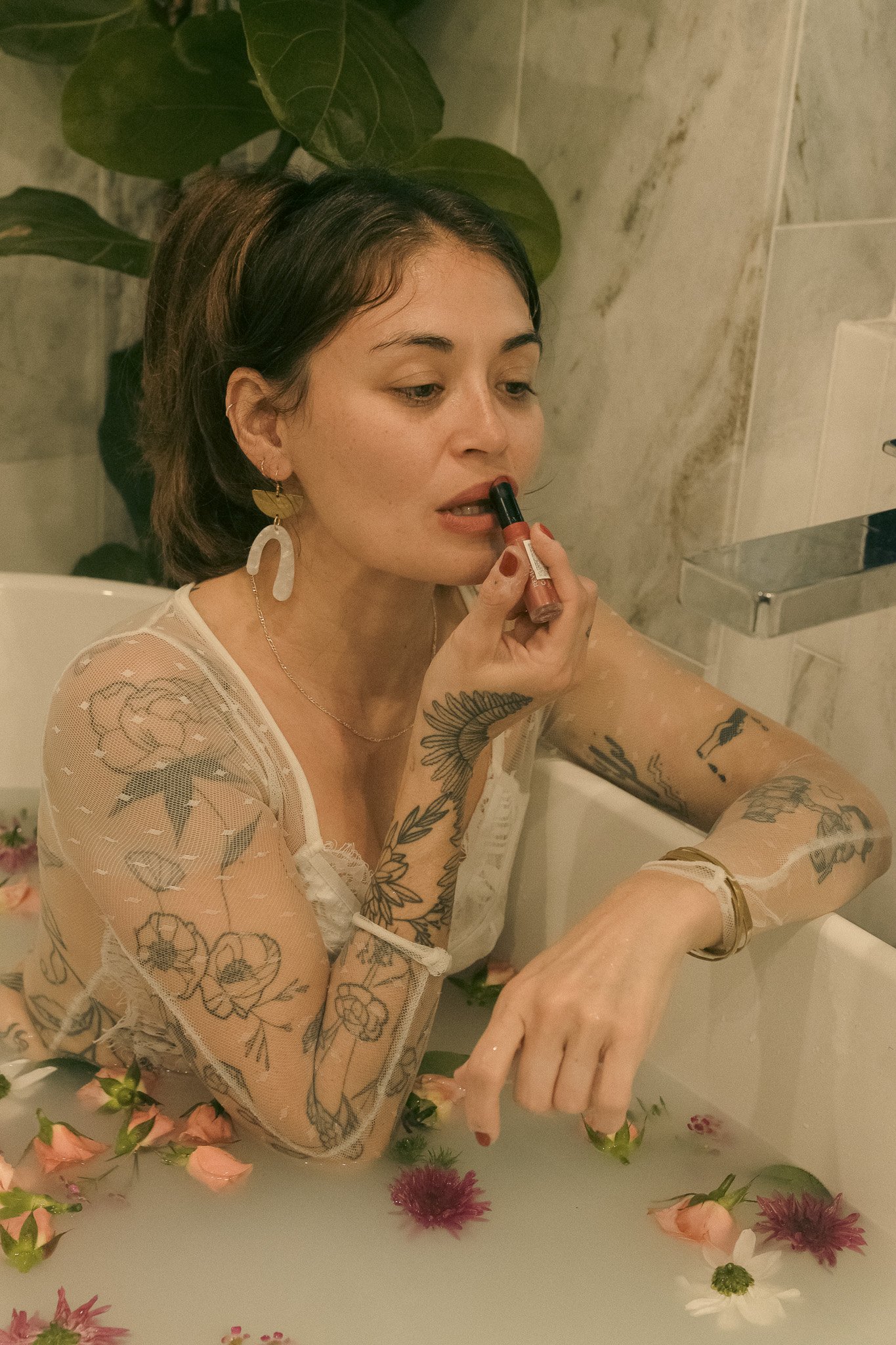 A woman with tattoos and lace clothing applying lipstick in a bathtub filled with milk and flowers.