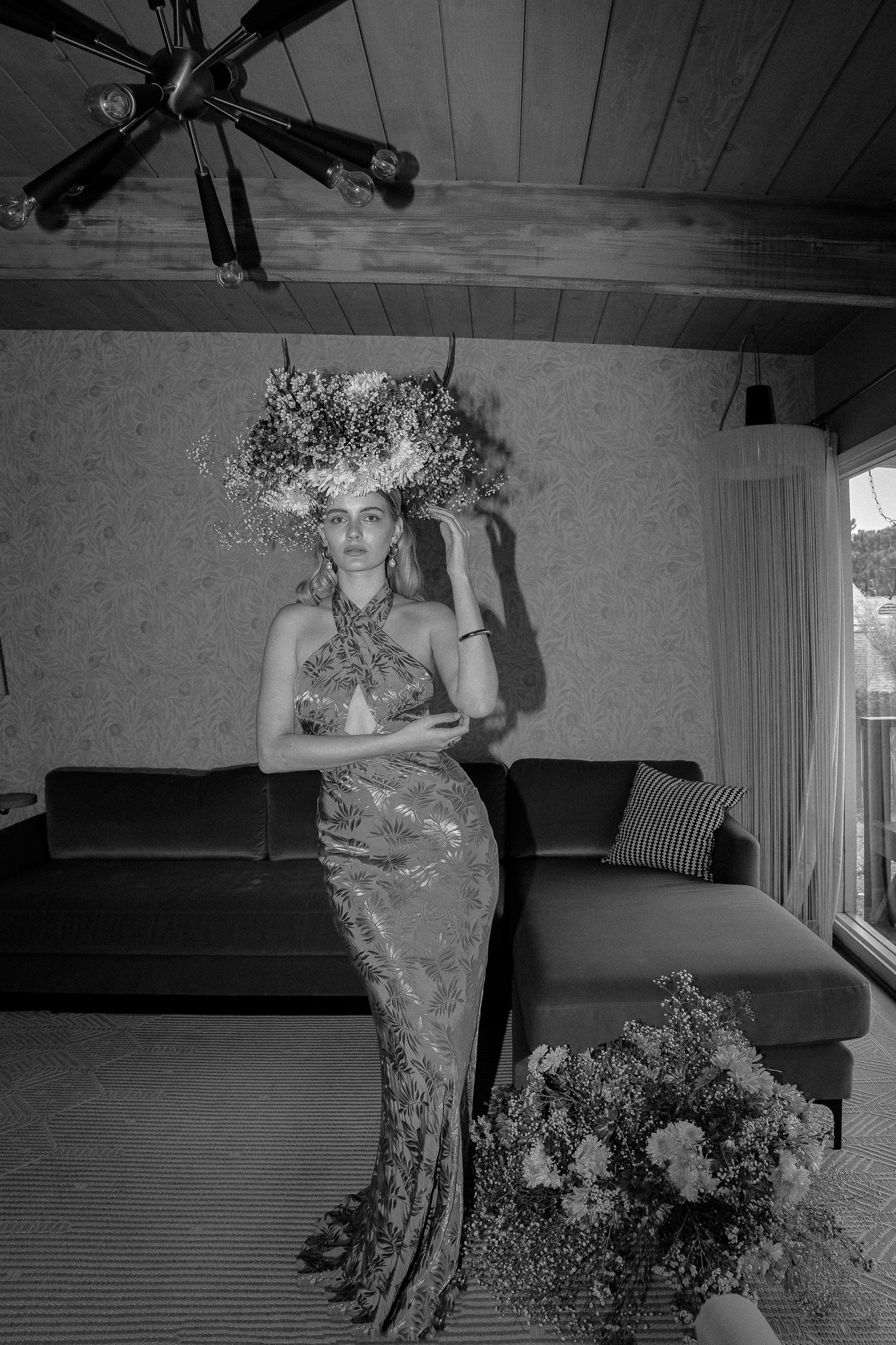Black and white photo of a woman in a floral dress and elaborate flower hat, standing in a room with a mid-century modern interior.