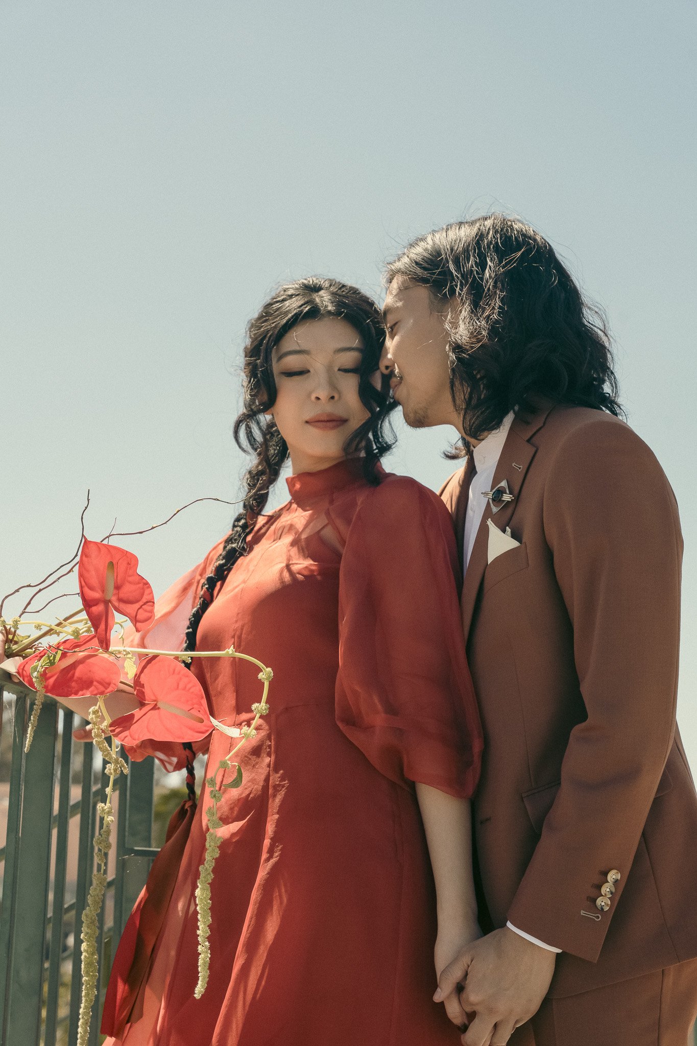 Couple in elegant attire holding hands outdoors, woman in red dress with braided hair, man in brown suit, red flowers in foreground.