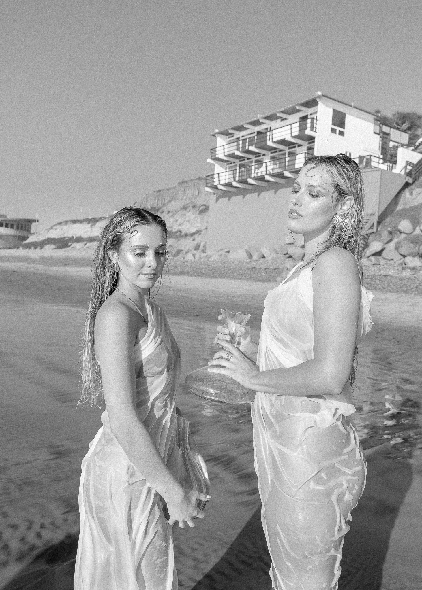 Two women in sheer dresses holding glass containers on a beach, with a modern building in the background.