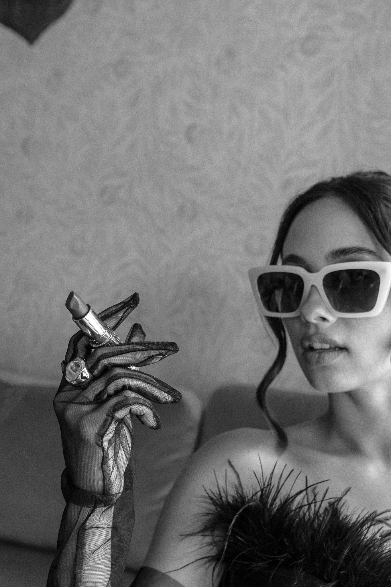 Black and white photo of a person wearing sunglasses and a feathered outfit, holding a lipstick in gloved hand.