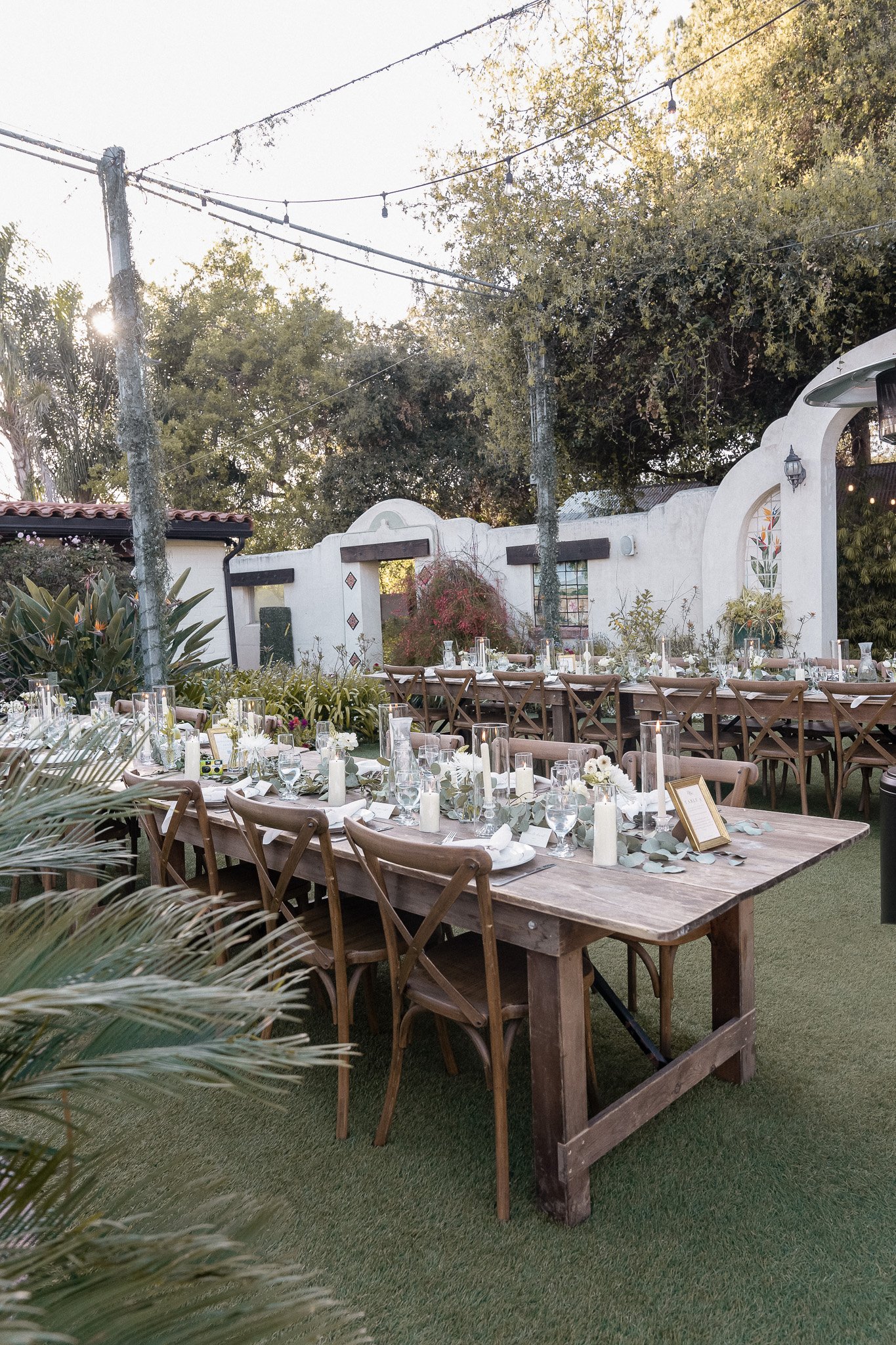 Outdoor garden event setup with wooden tables and chairs, decorated with candles, greenery, and flowers, under string lights.