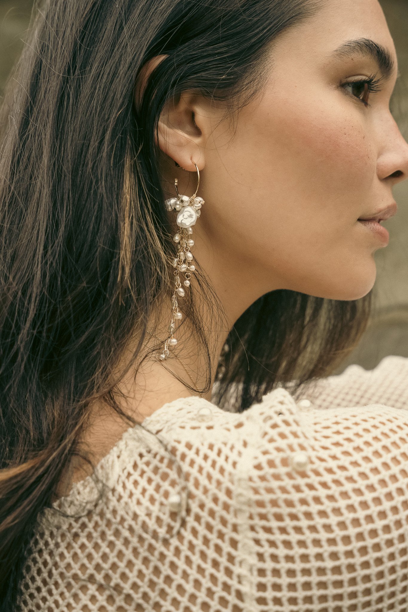 Close-up of a woman wearing a long, ornate earring with beads and pearls, and a white crocheted top.