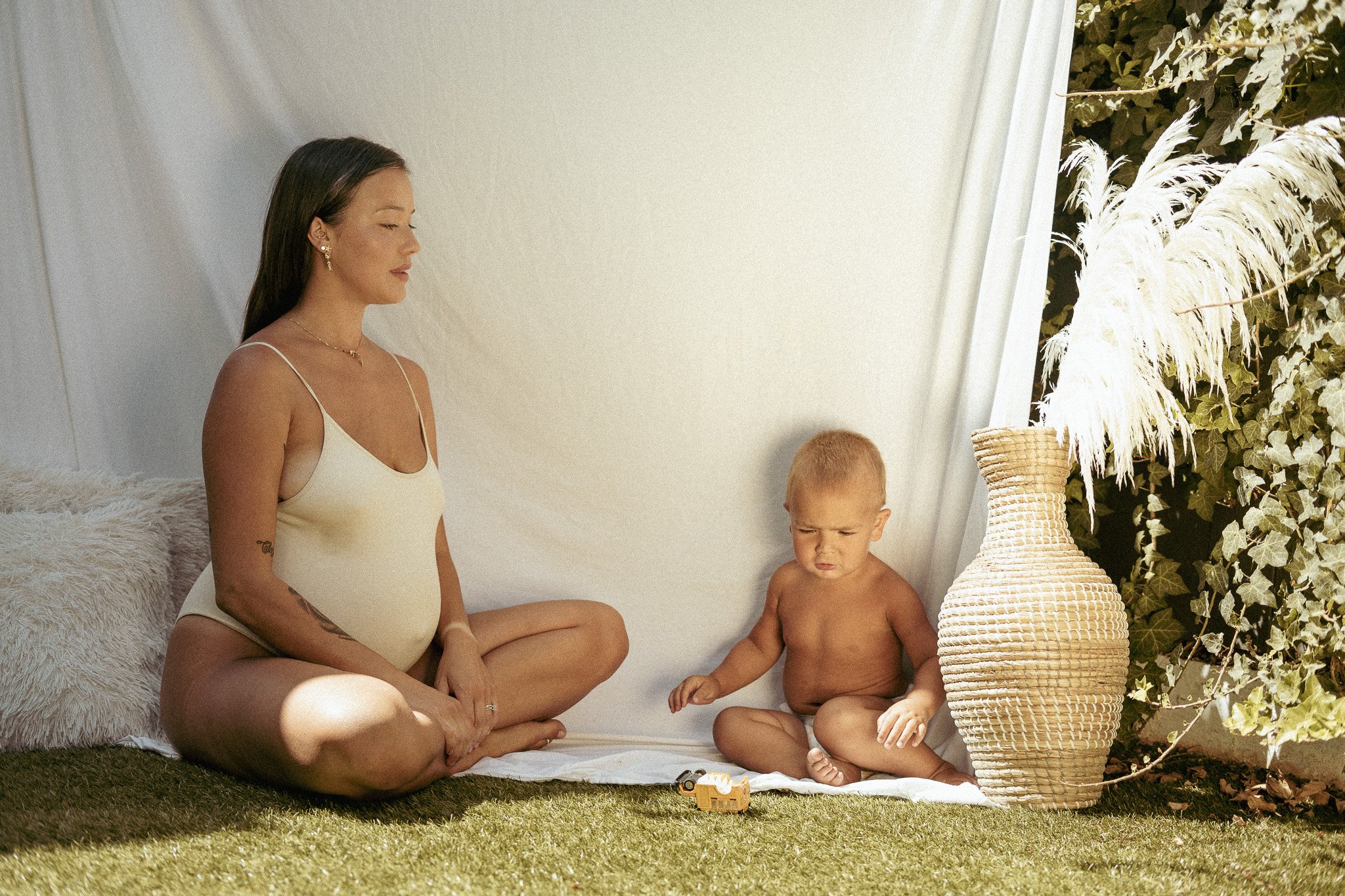 Pregnant woman sitting beside a baby who is playing, with a large woven vase nearby, outdoors against a white draped backdrop.