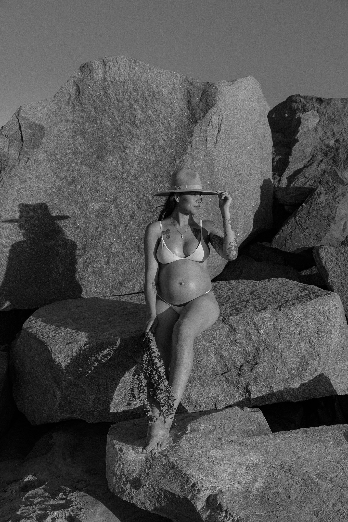 Pregnant woman in bikini and hat sitting on large rocks, monochrome photo.