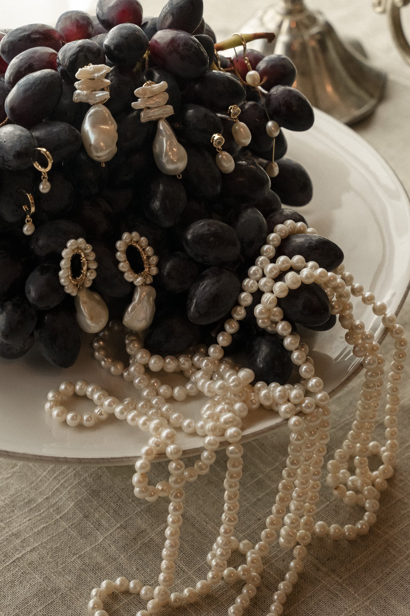 Bunch of black grapes on a plate adorned with pearl necklaces and earrings.