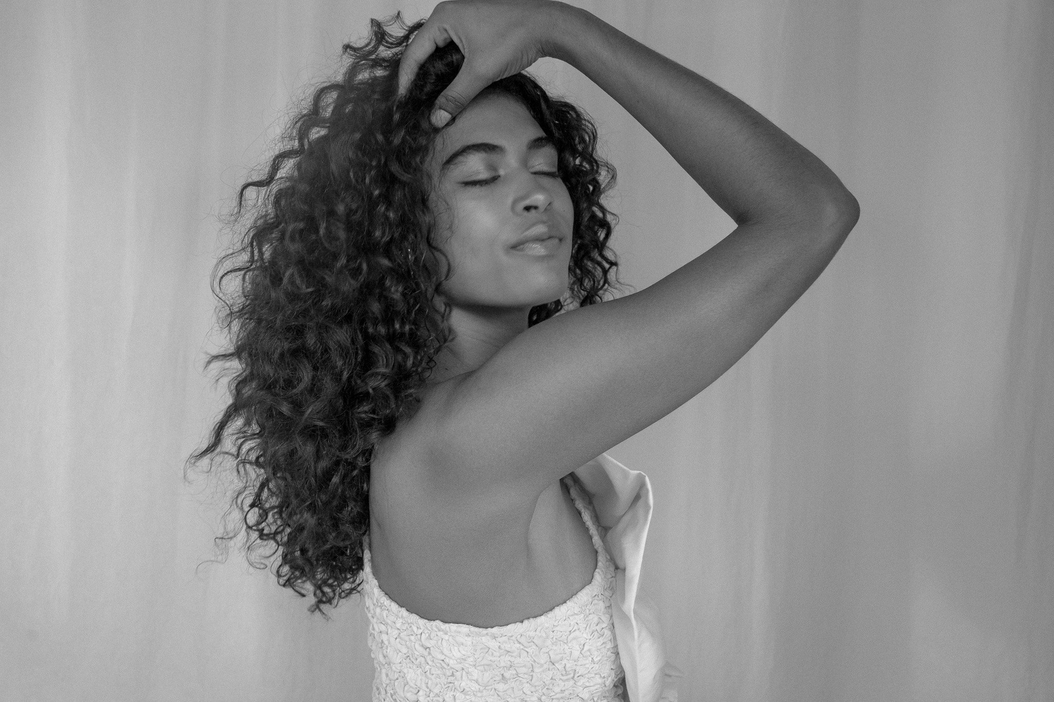 Black and white photo of a person with long curly hair wearing a textured, strapless top, with one arm raised and eyes closed.