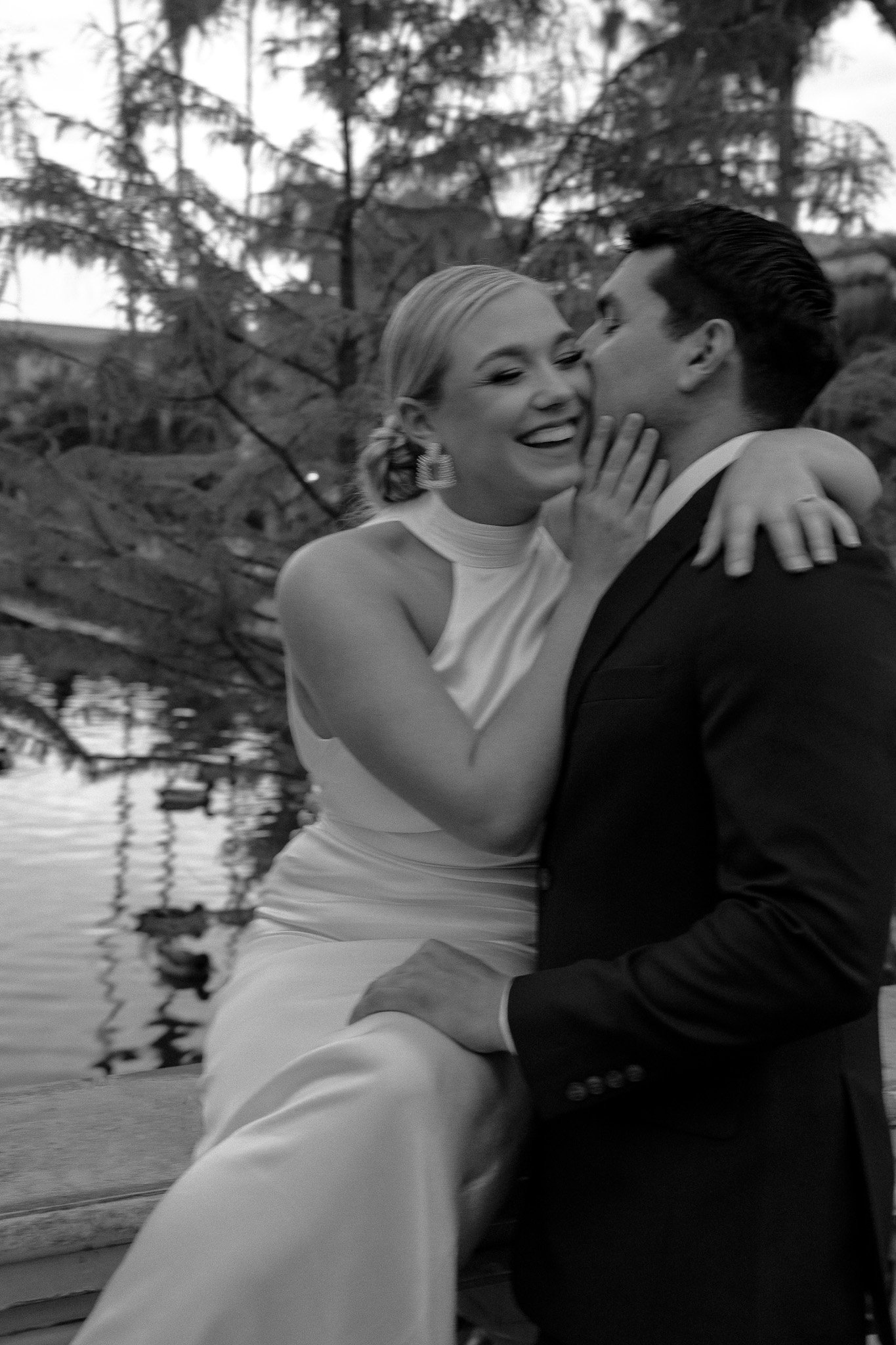 Black and white photo of a smiling couple, with the woman in a sleeveless dress and the man in a suit, embracing near a pond with trees in the background.