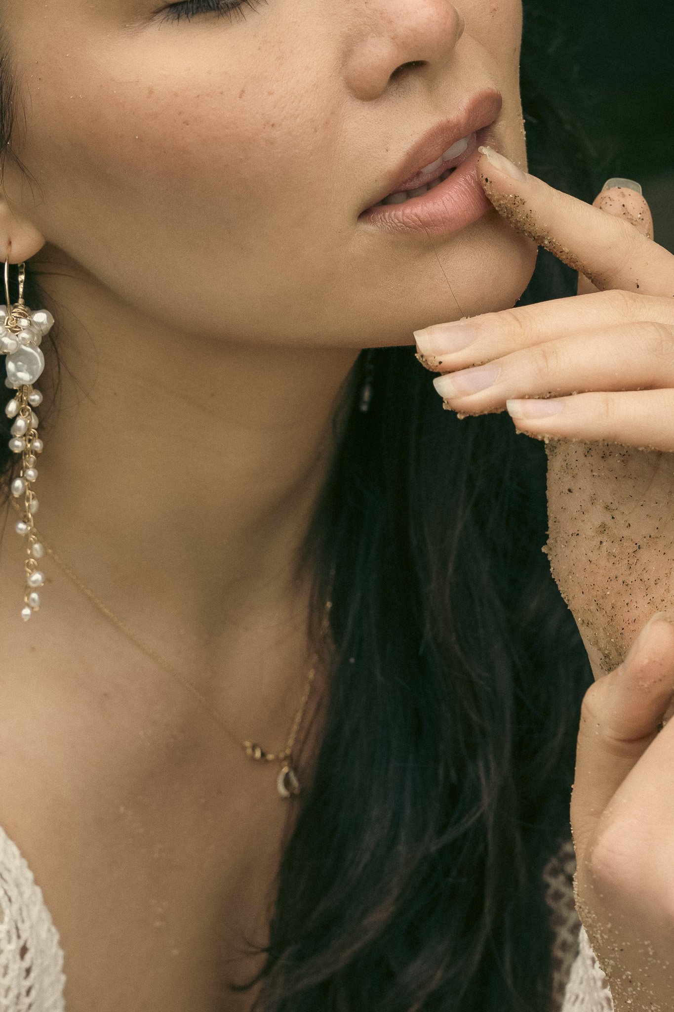 Close-up of a woman's face with sand on her lips and fingers, wearing a pearl earring and necklace.